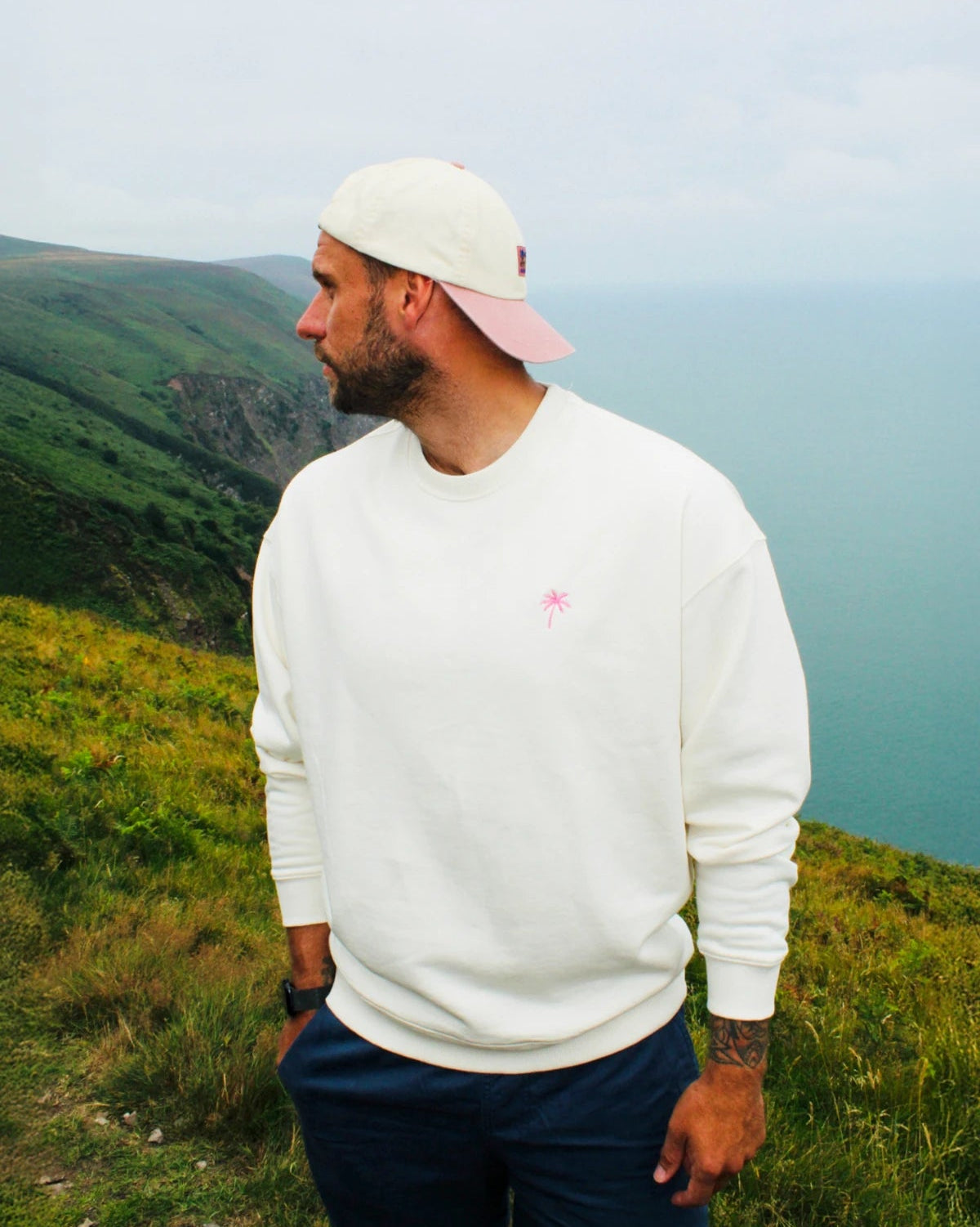 Model trägt den Palm Tree Oversized Sweater Cream in Kombination mit Shorts und Cap bei einer Wanderung an der Küste.