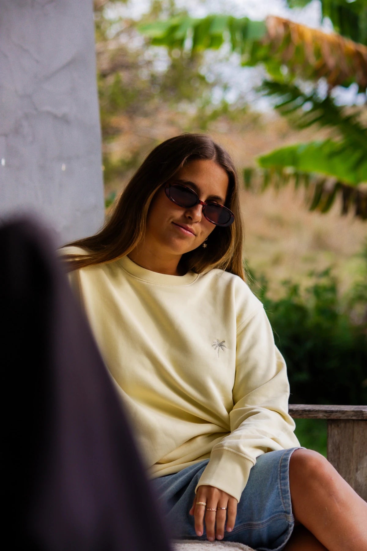 Model sitzt lässig auf Holzbank in buttergelbem Sweater und Jeansshorts – The Sunkissed Rebels Vibes pur.