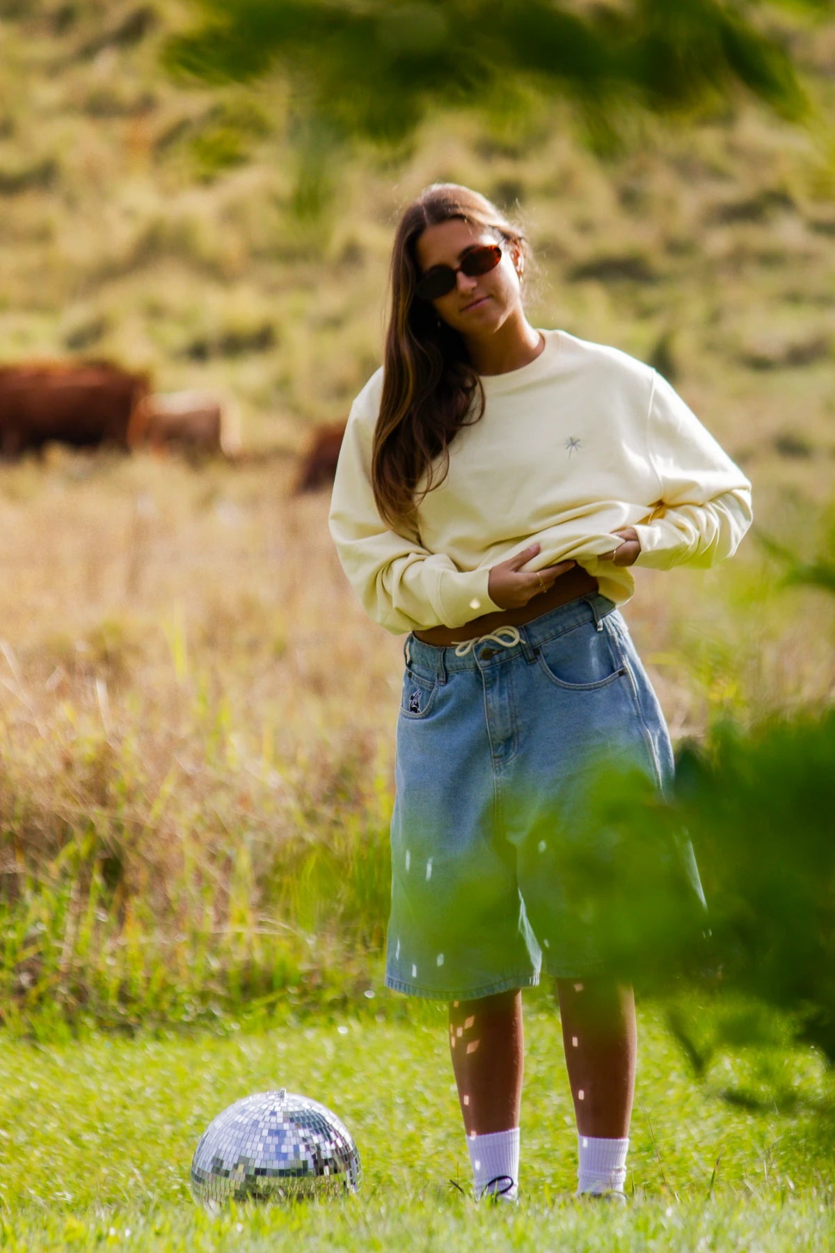 Model steht auf Wiese mit Discokugel im butterfarbenen Pullover und Denim Shorts – Palmenstick vorne sichtbar.