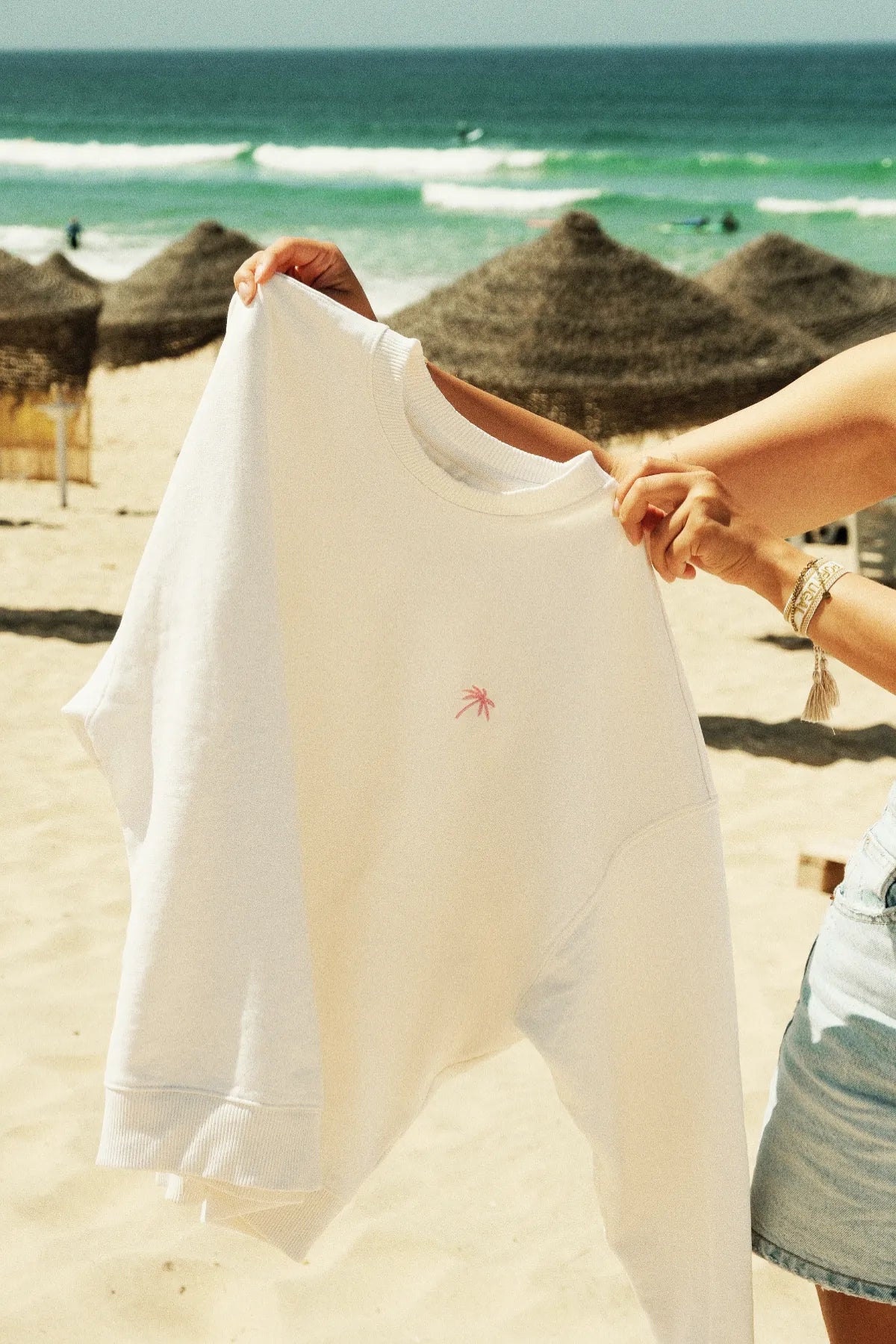 Palm Tree Oversized Sweater Cream – gehalten vor Meereskulisse mit Sonnenschirmen