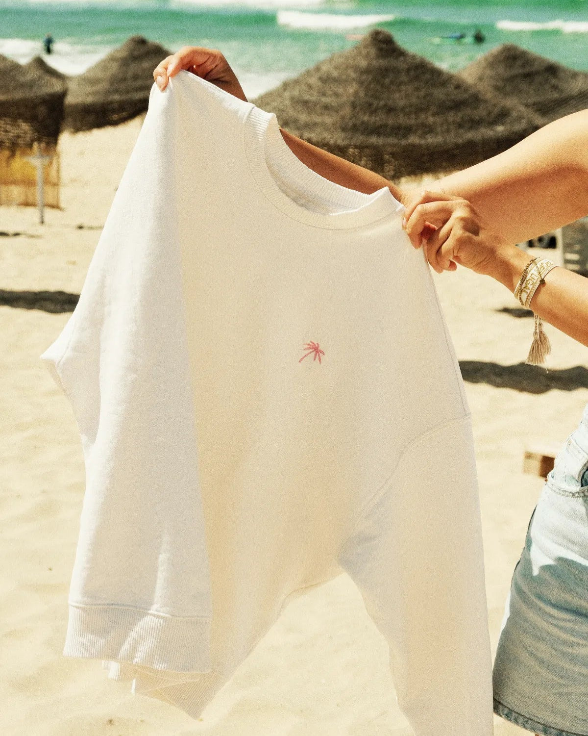 Palm Tree Oversized Sweater Cream – gehalten vor Meereskulisse mit Sonnenschirmen