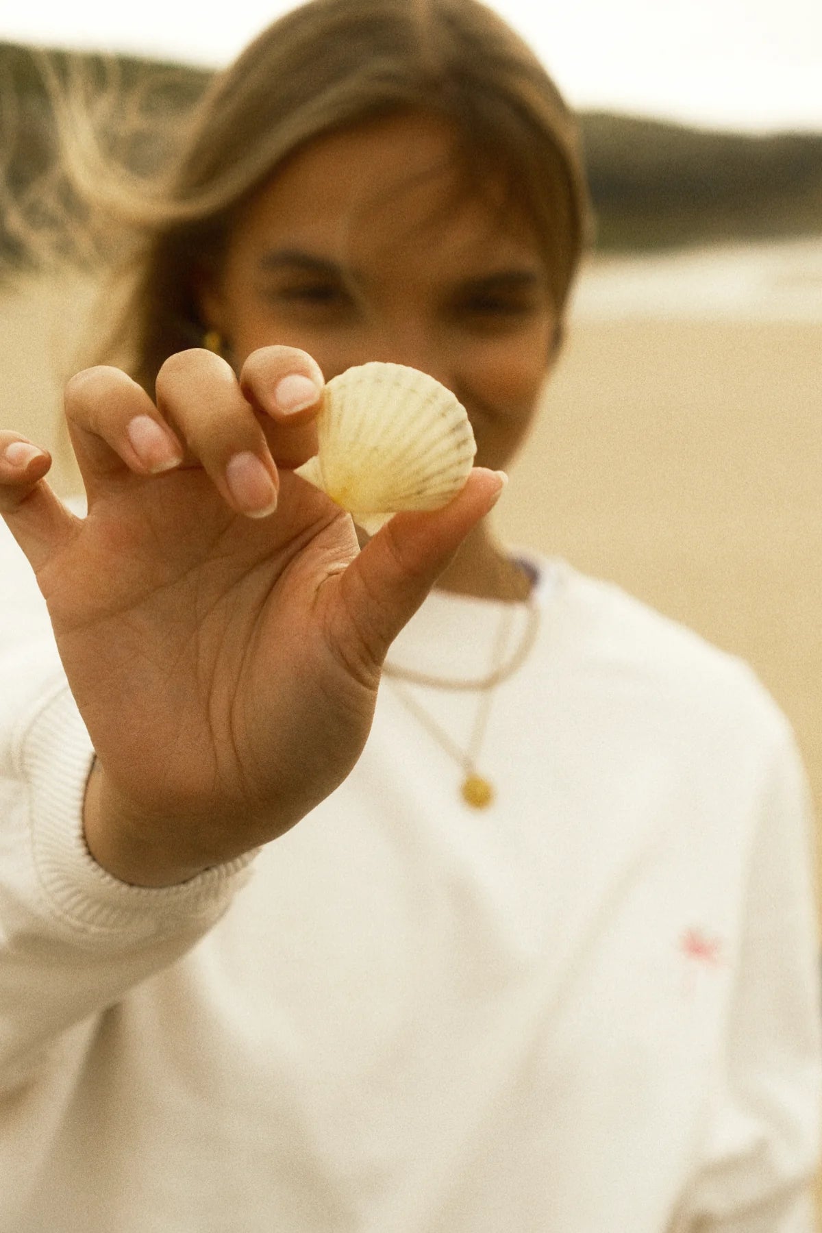 Model im weißen Sweater zeigt eine Muschel am Strand in sommerlicher Stimmung.