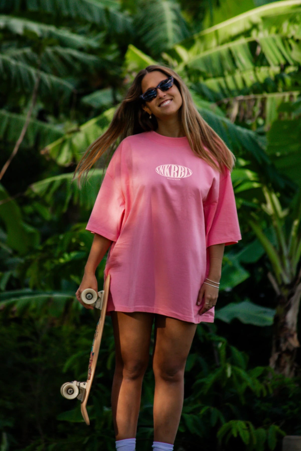 Lächelnde Skaterin in tropischer Kulisse trägt das pinke SNKRBLS Oversized T-Shirt in Pink Joy mit Sonnenbrille und Skateboard – urbaner Sommerstyle pur