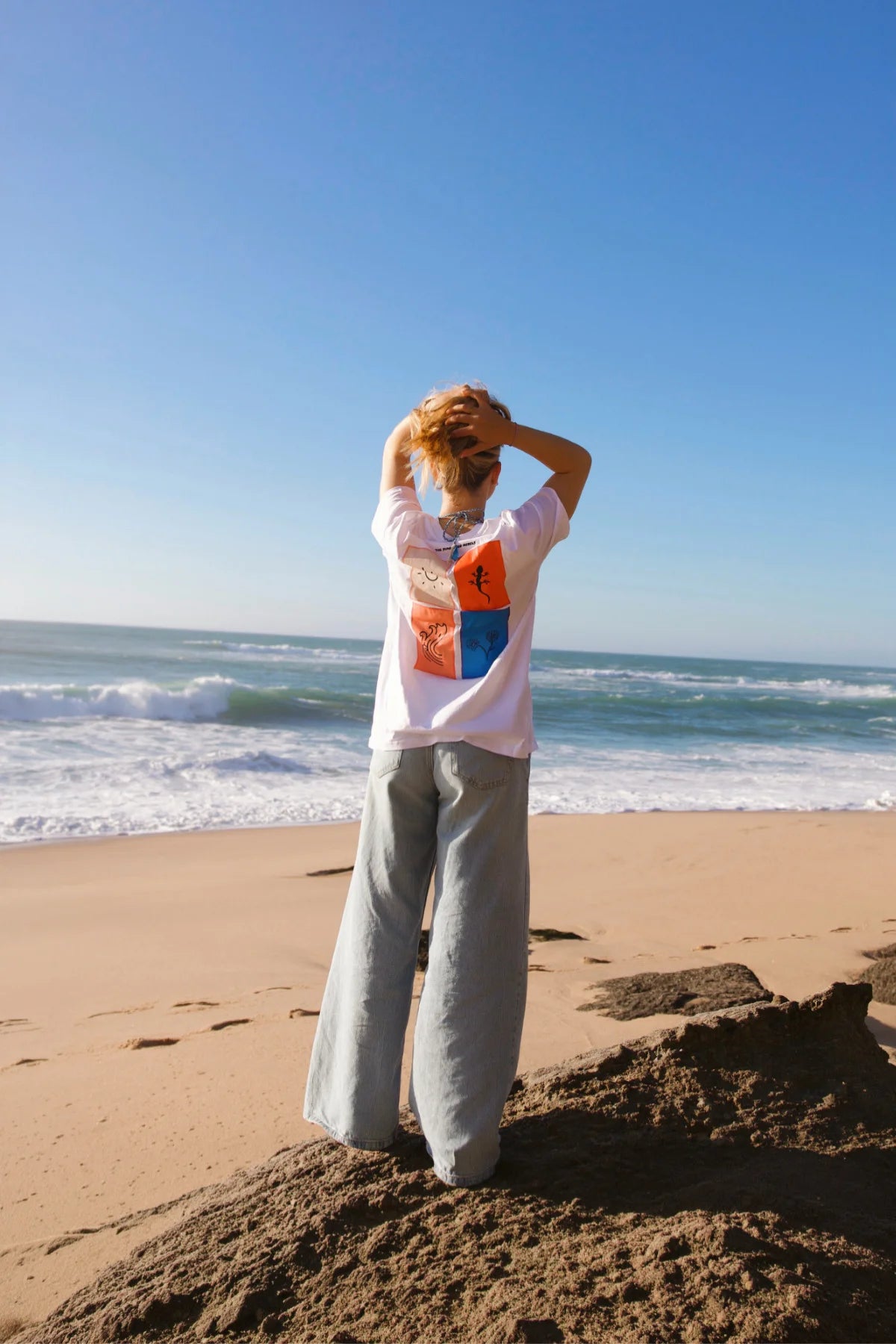 ückansicht des weißen Rebel Elements Light T-Shirts am Strand; Fokus auf den großen vierteiligen Rückenprint mit Sonne, Gecko, Welle und Blumen-Motiv.