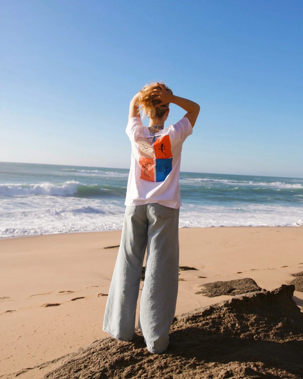 ückansicht des weißen Rebel Elements Light T-Shirts am Strand; Fokus auf den großen vierteiligen Rückenprint mit Sonne, Gecko, Welle und Blumen-Motiv.