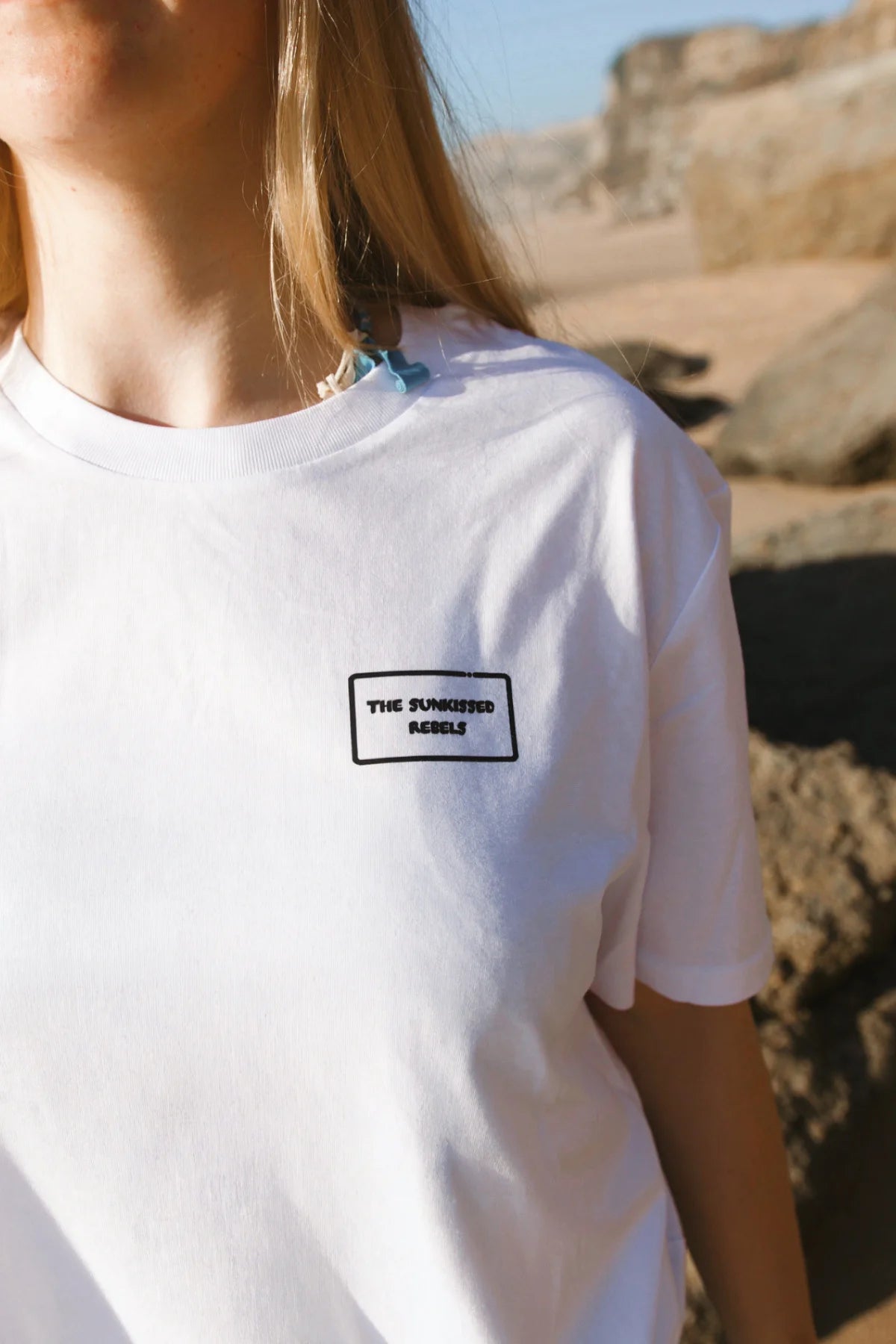 Nahaufnahme der Vorderseite des weißen Light T-Shirts mit dem dezenten, schwarz eingerahmten „The Sunkissed Rebels“ Logo-Patch auf der Brust.