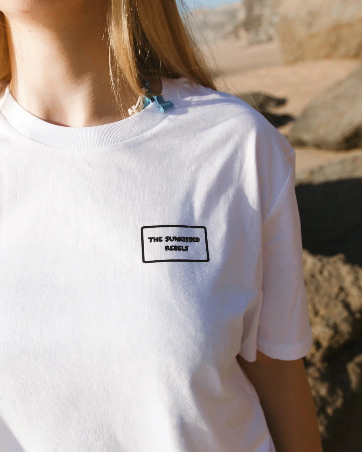 Nahaufnahme der Vorderseite des weißen Light T-Shirts mit dem dezenten, schwarz eingerahmten „The Sunkissed Rebels“ Logo-Patch auf der Brust.