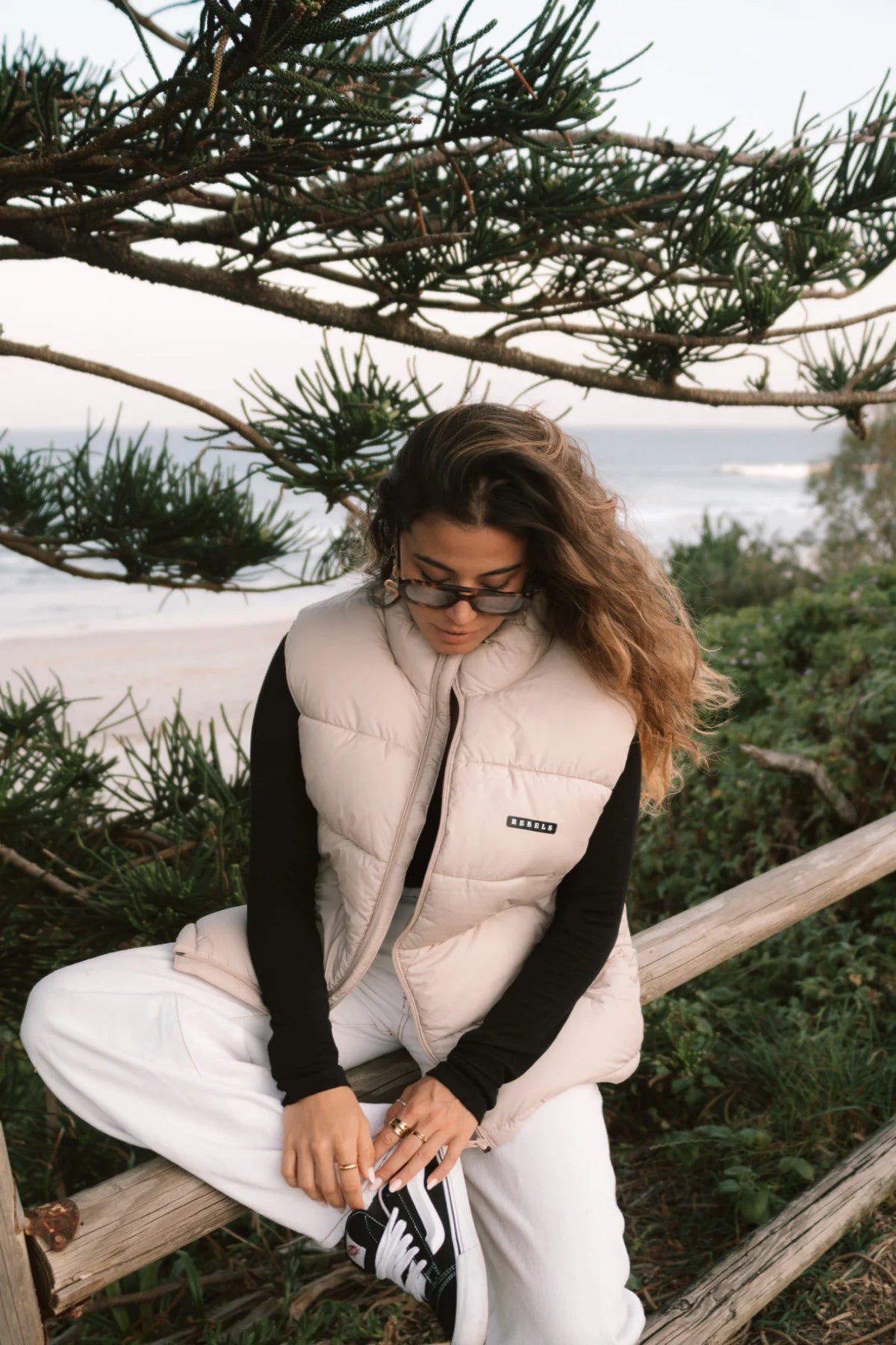 Entspannte Szene am Strand – Frau sitzt im Rebels Puffer Outdoor Vest Beige von The Sunkissed Rebels auf einem Holzgeländer.