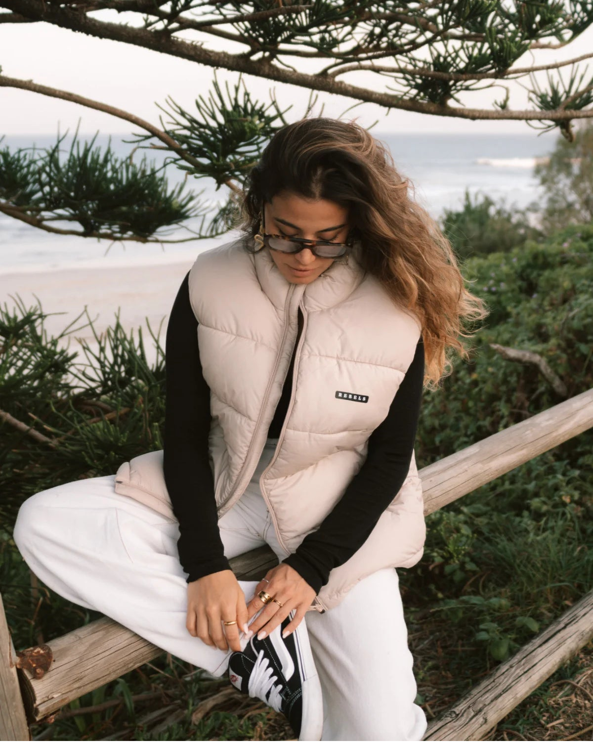Entspannte Szene am Strand – Frau sitzt im Rebels Puffer Outdoor Vest Beige von The Sunkissed Rebels auf einem Holzgeländer.