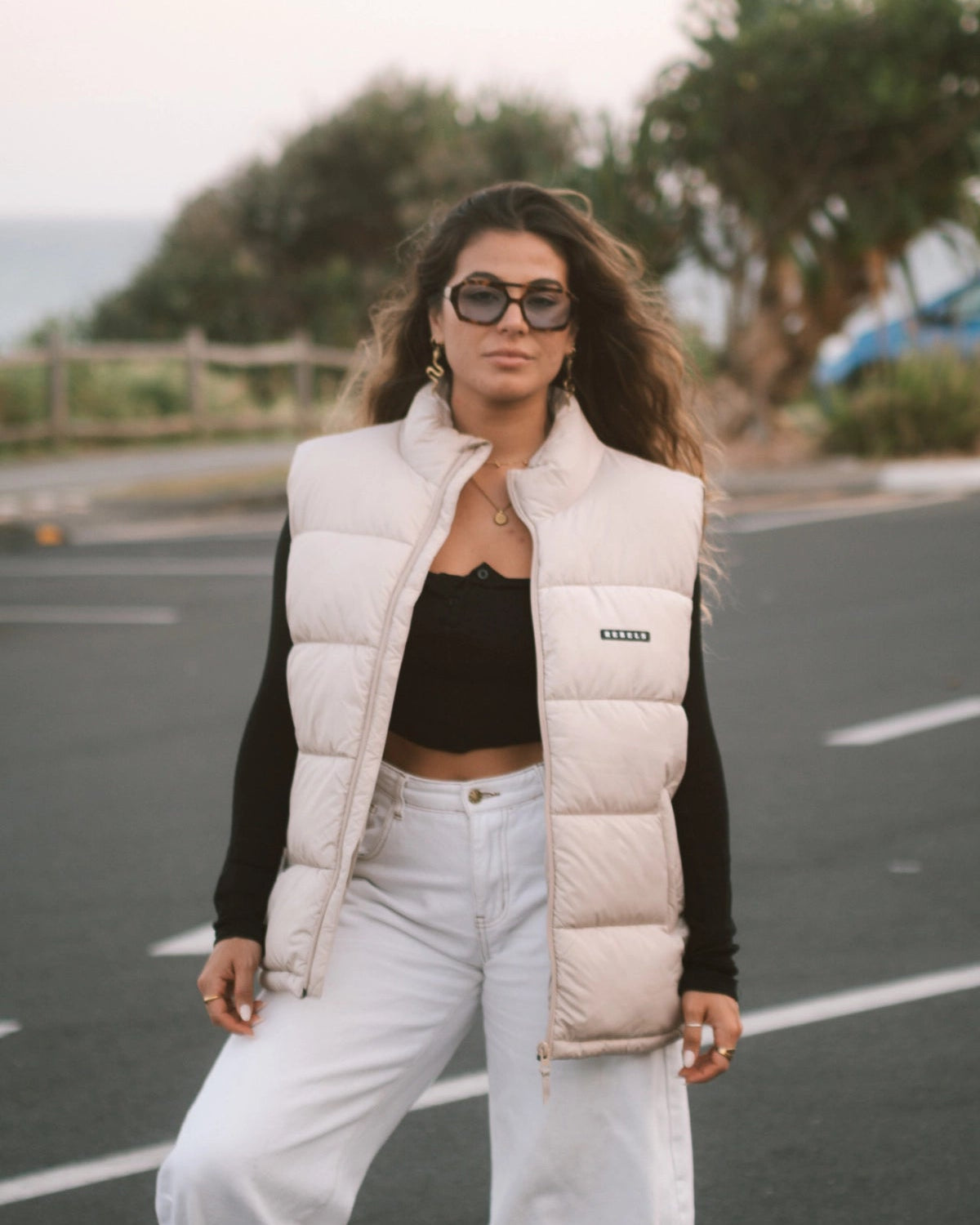 Streetstyle-Look – Frau mit der Rebels Puffer Outdoor Vest Beige von The Sunkissed Rebels steht auf einem Parkplatz und schaut in Kamera
