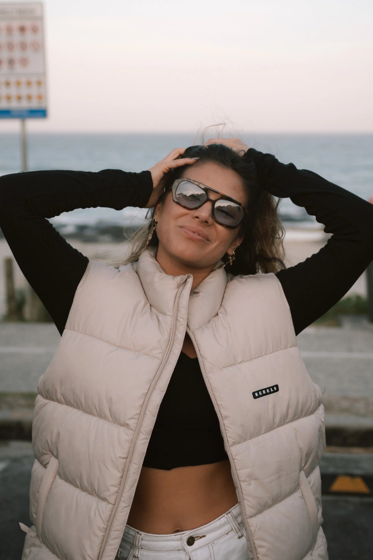 Frau trägt die Rebels Puffer Outdoor Vest in Beige von The Sunkissed Rebels, lässig kombiniert mit schwarzem Longsleeve und weißer Hose am Strand.