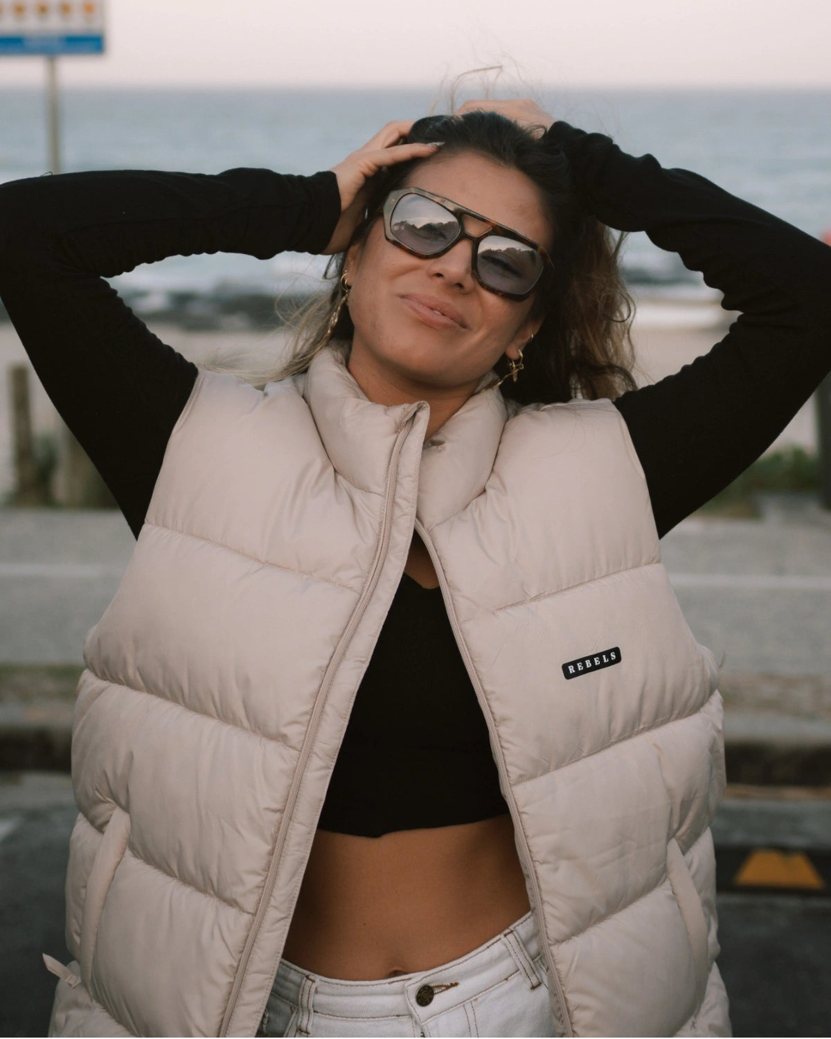 Frau trägt die Rebels Puffer Outdoor Vest in Beige von The Sunkissed Rebels, lässig kombiniert mit schwarzem Longsleeve und weißer Hose am Strand.