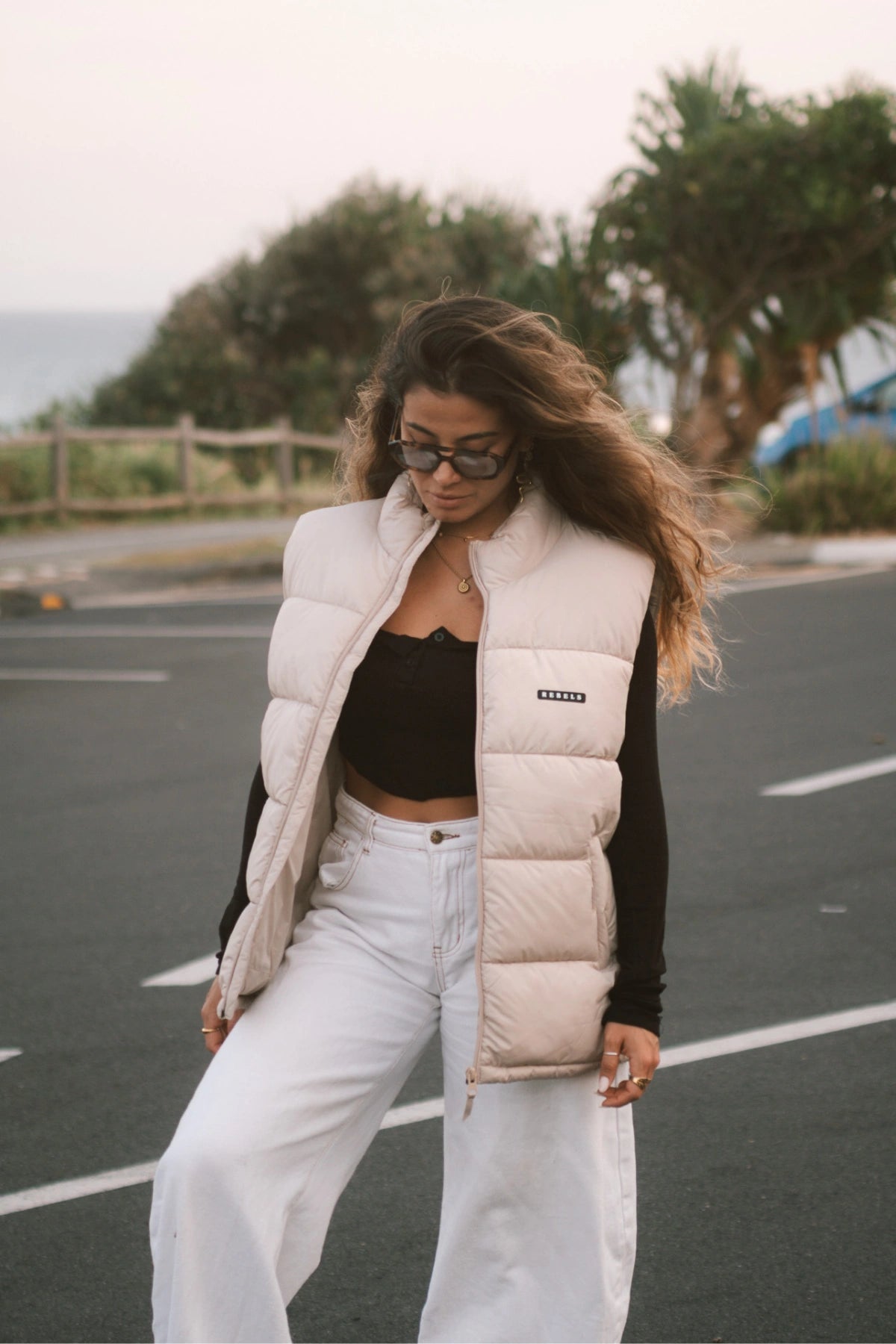 Streetstyle-Look – Frau mit der Rebels Puffer Outdoor Vest Beige von The Sunkissed Rebels steht auf einem Parkplatz mit Meerblick.