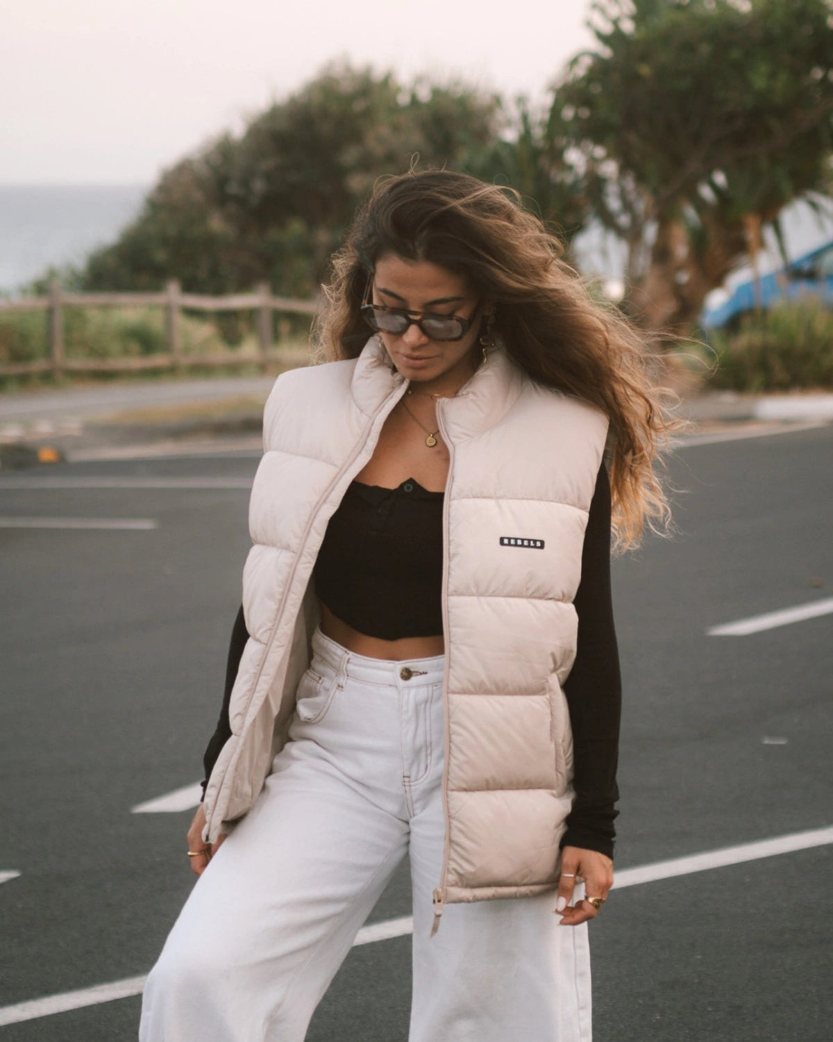 Streetstyle-Look – Frau mit der Rebels Puffer Outdoor Vest Beige von The Sunkissed Rebels steht auf einem Parkplatz mit Meerblick.