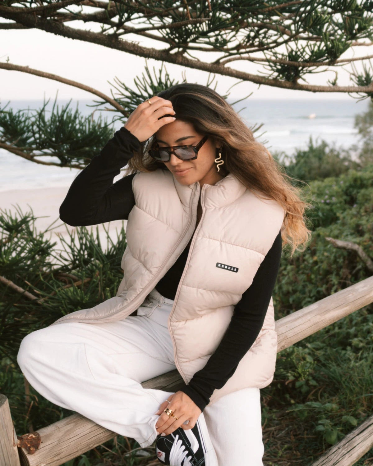 Streetstyle-Look – Frau mit der Rebels Puffer Outdoor Vest Beige von The Sunkissed Rebels sitzt auf Holzbalken am Strand