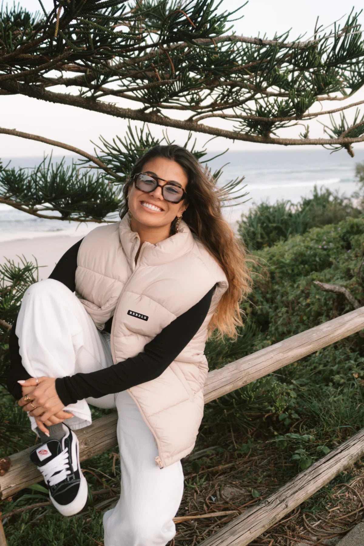 Frau mit der Rebels Puffer Outdoor Vest Beige von The Sunkissed Rebels – modischer Herbst-Look am Meer mit weitem Hosenstil.