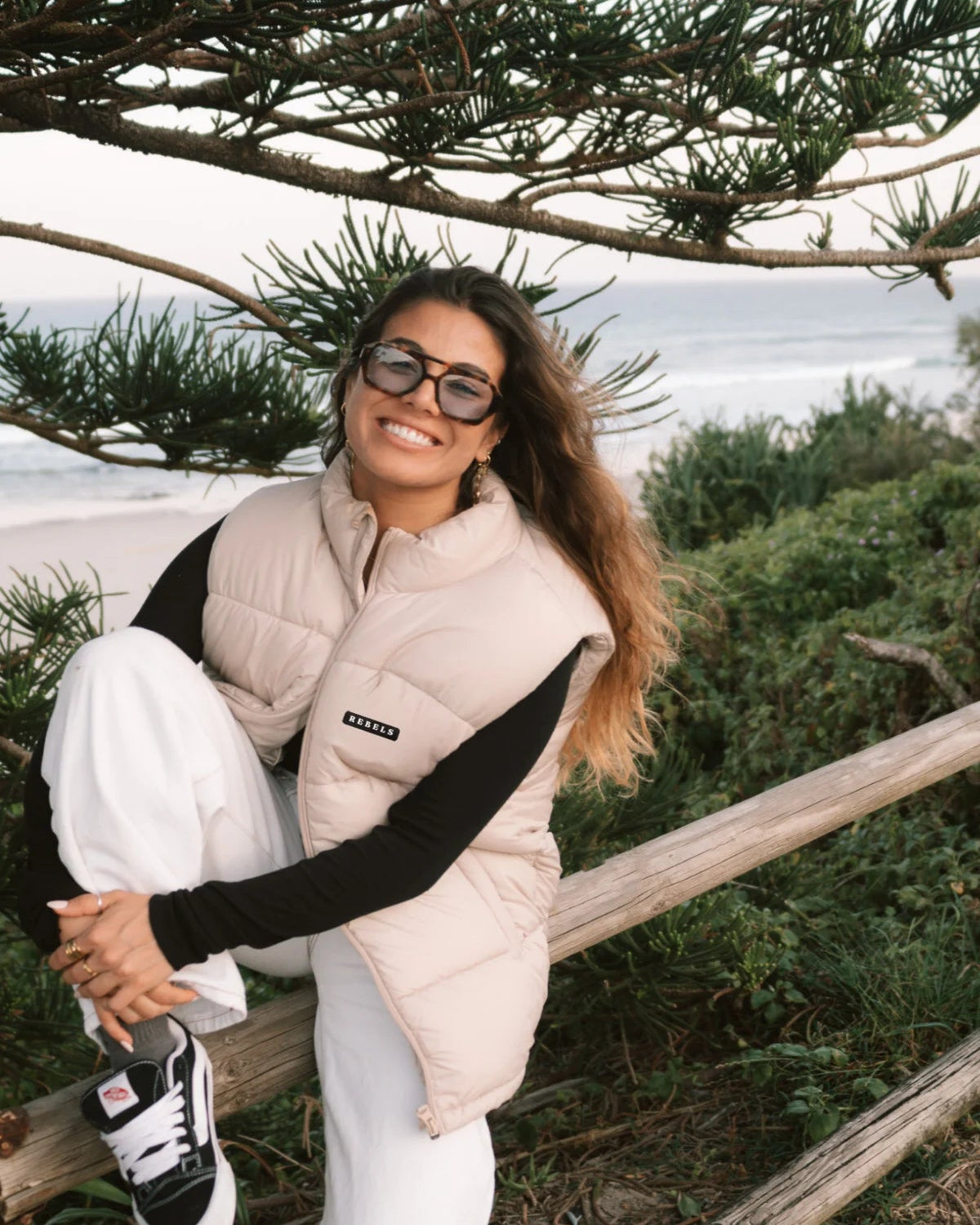 Frau mit der Rebels Puffer Outdoor Vest Beige von The Sunkissed Rebels – modischer Herbst-Look am Meer mit weitem Hosenstil.