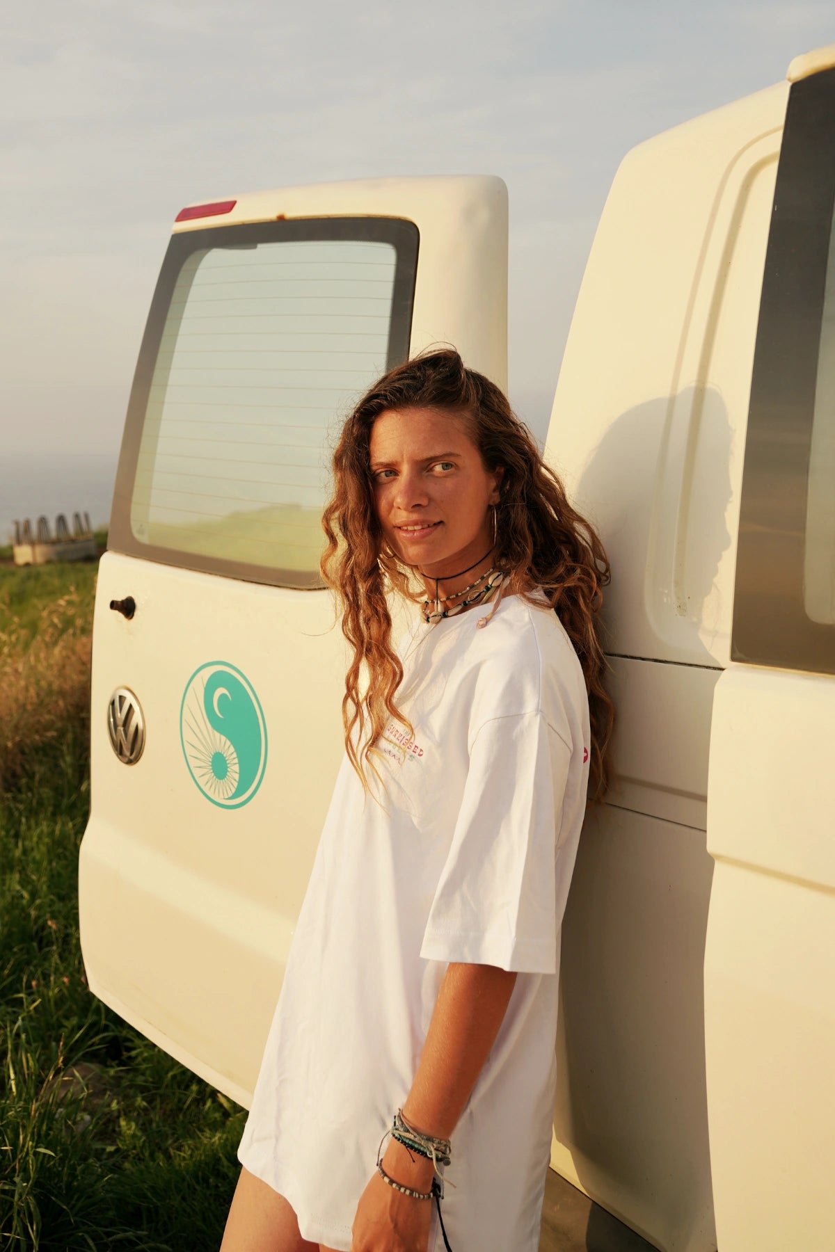 Frau mit Boho-Schmuck im weißen Vacation Vibes Shirt lehnt lässig an VW Camper im Abendlicht