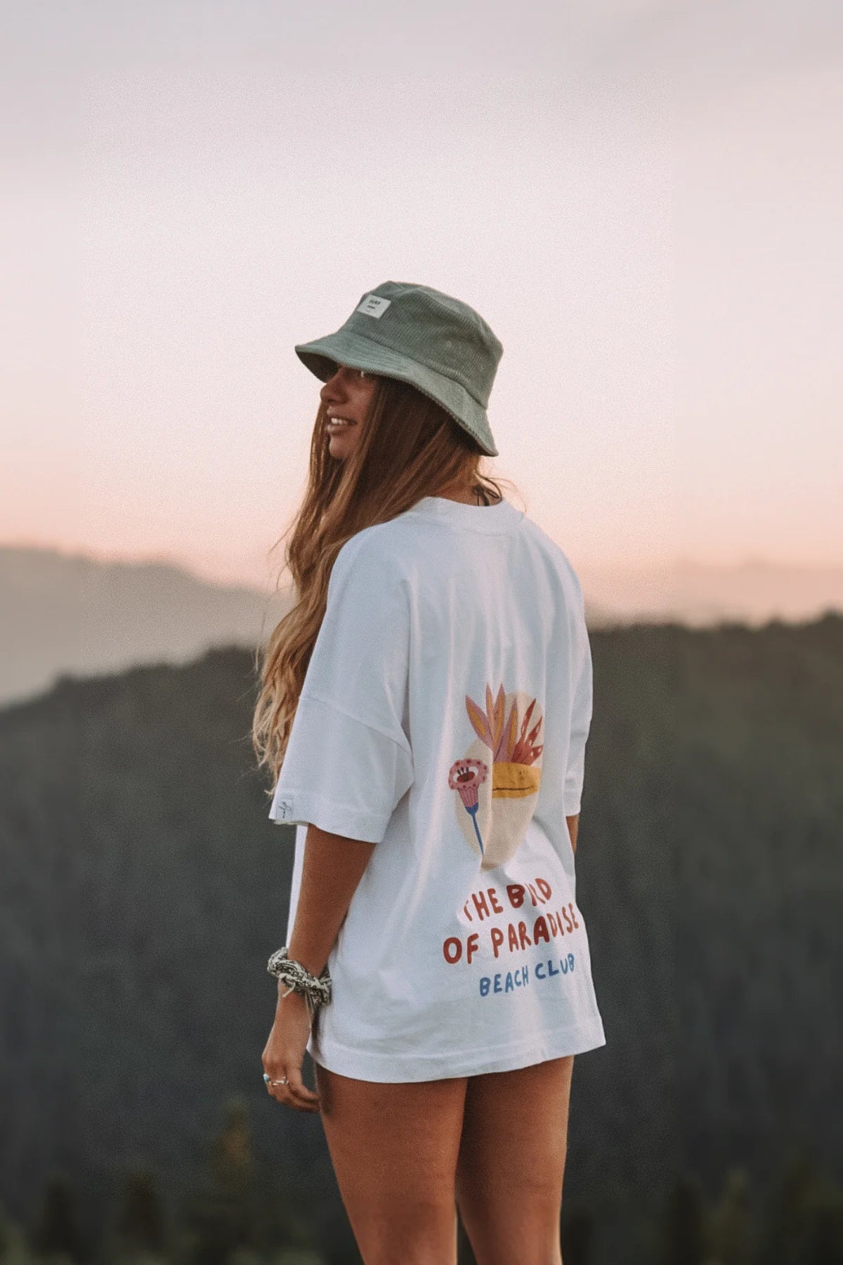 Weißes Oversized T-Shirt The Bird of Paradise von The Sunkissed Rebels – Rückenansicht bei Sonnenuntergang in den Bergen mit tropischem Print