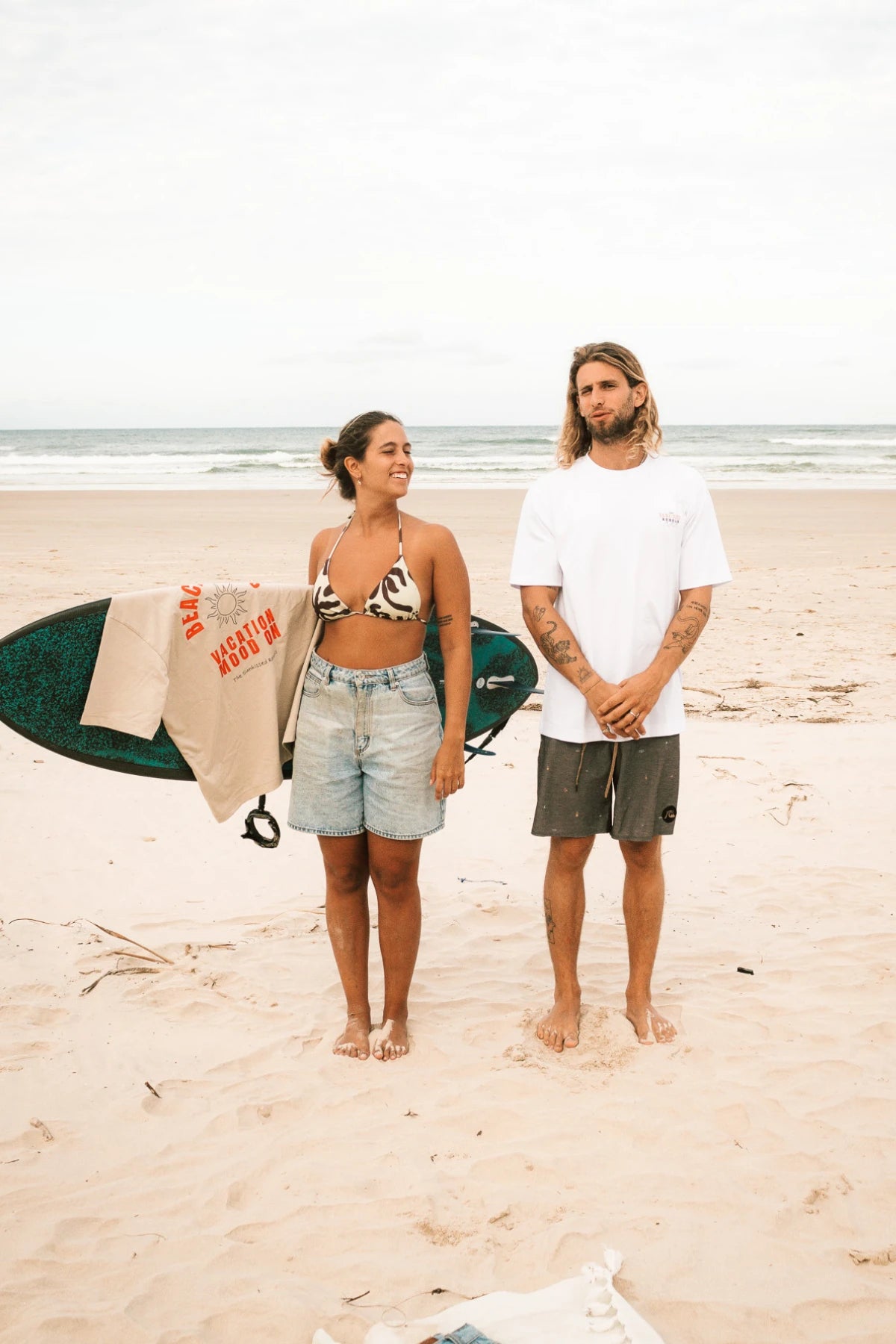 Mann und Frau frontal am Strand, er trägt Paradise-&-Sunset-Shirt, sie hält ein Surfbrett