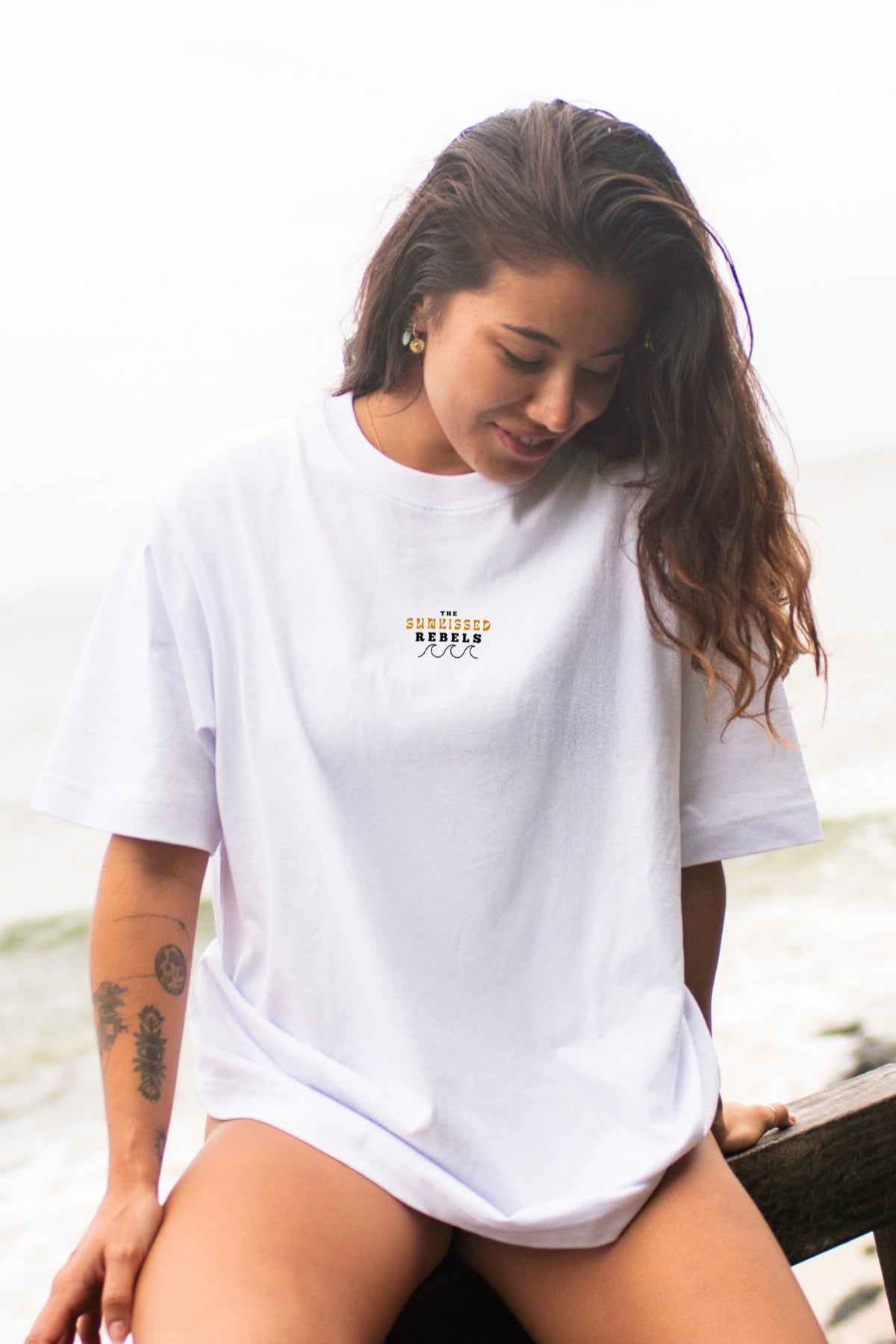 Lächelnde Frau im weißen Oversized T-Shirt Paradise & Sunset von The Sunkissed Rebels vor Ozeankulisse