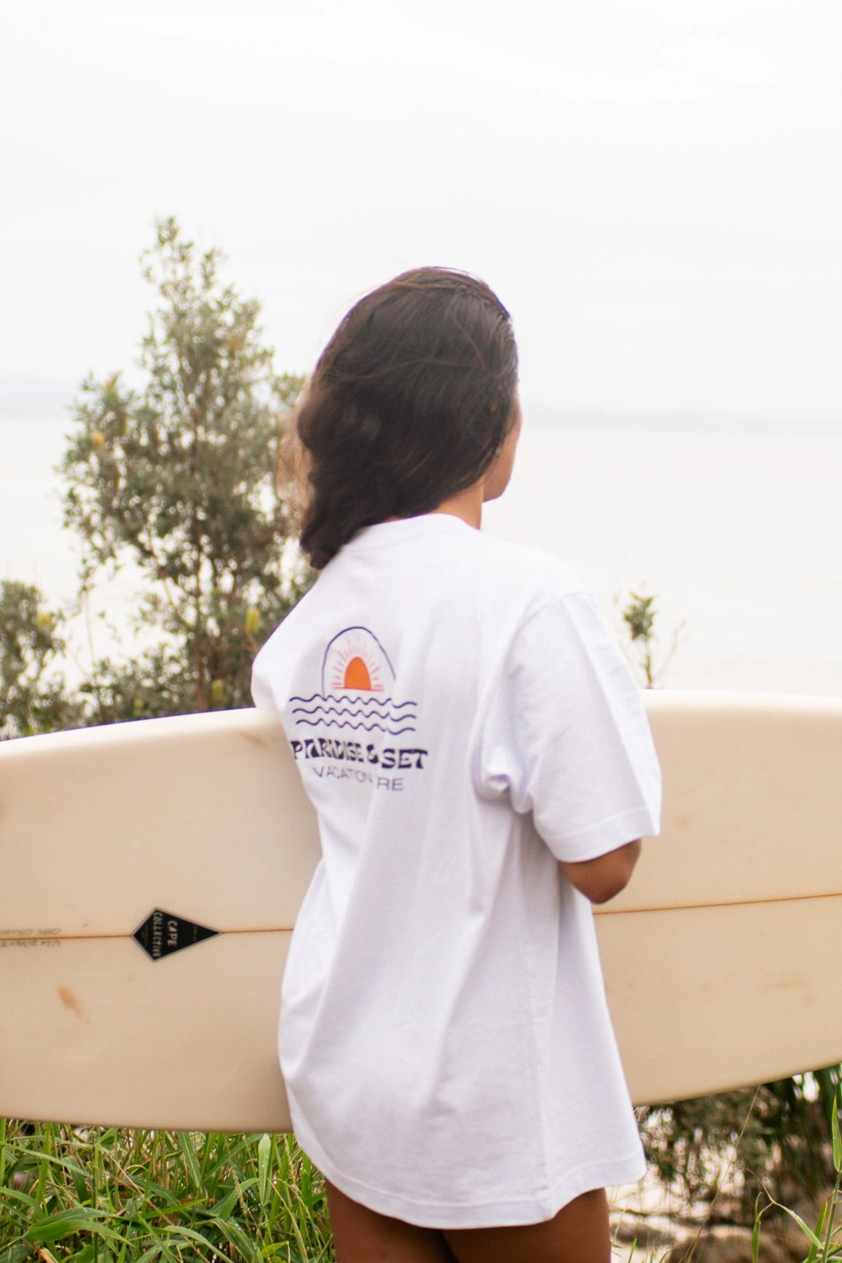 Rückenansicht einer Frau im weißen Oversized T-Shirt Paradise & Sunset von The Sunkissed Rebels am Meer
