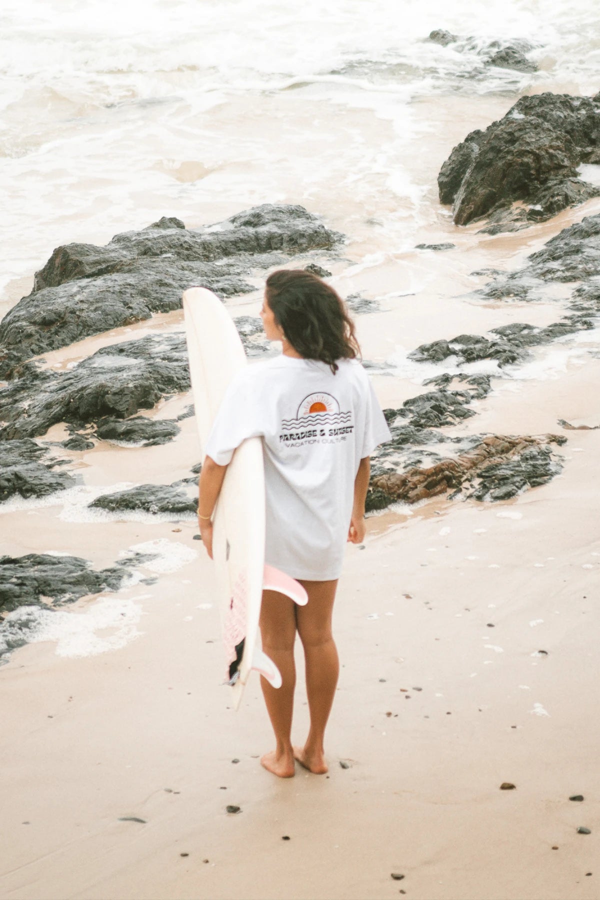 Frau mit Surfboard am Felsenstrand trägt weißes Oversized T-Shirt Paradise & Sunset von The Sunkissed Rebels