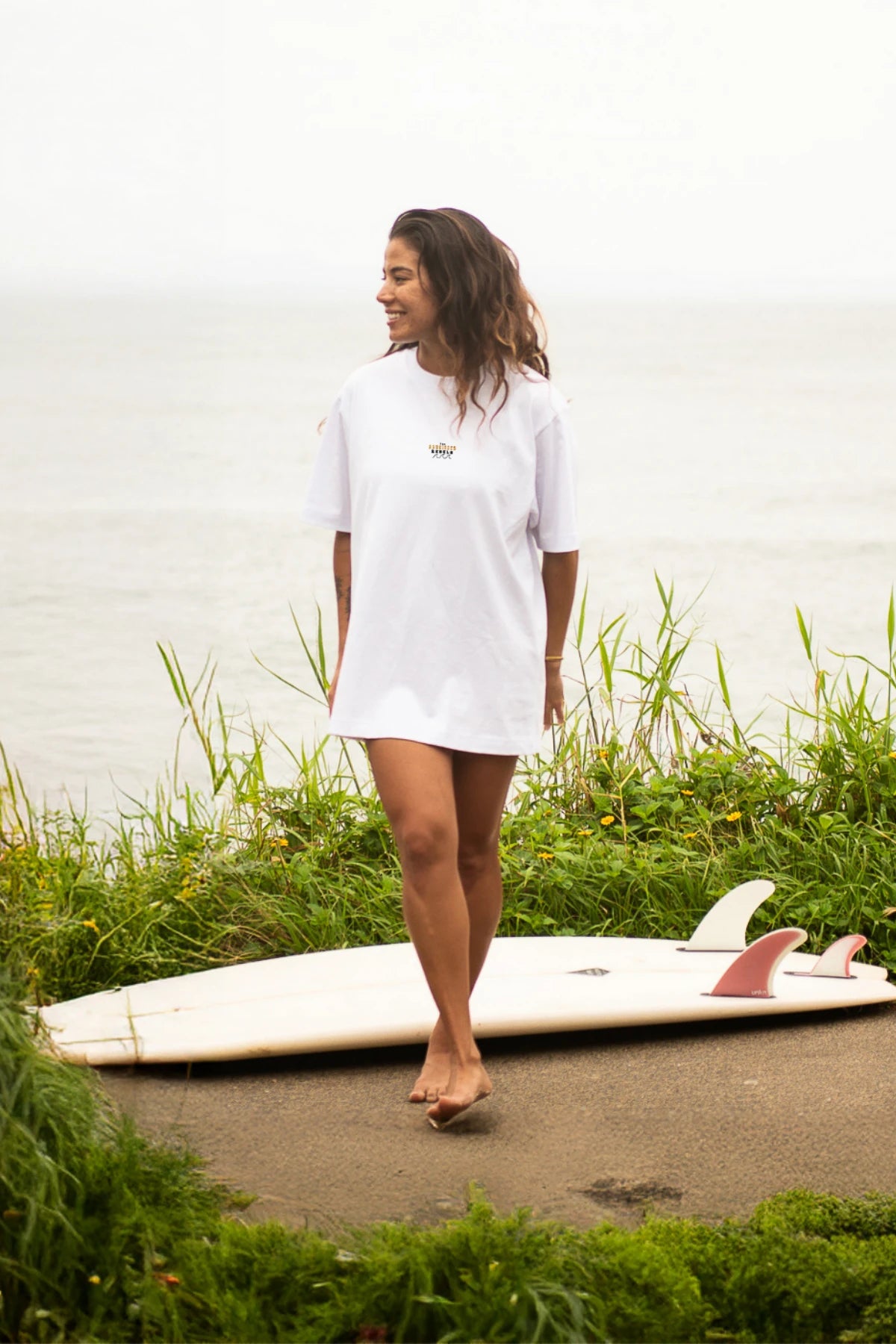 Junge Frau mit langen Haaren trägt weißes Oversized T-Shirt von The Sunkissed Rebels und steht vor einem Surfbrett