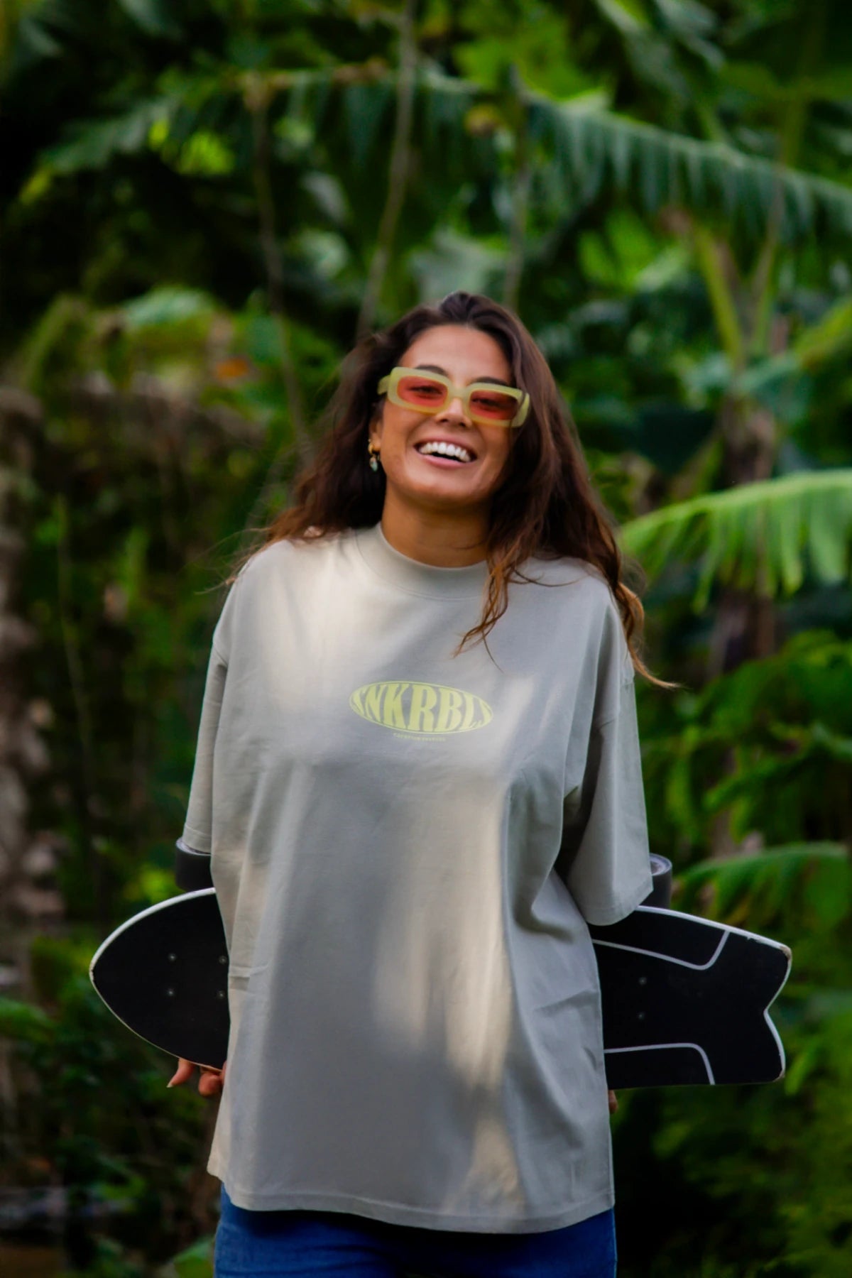 Frau im SNKRBLS Oversized T-Shirt in Stone steht lachend mit Skateboard vor tropischer Kulisse