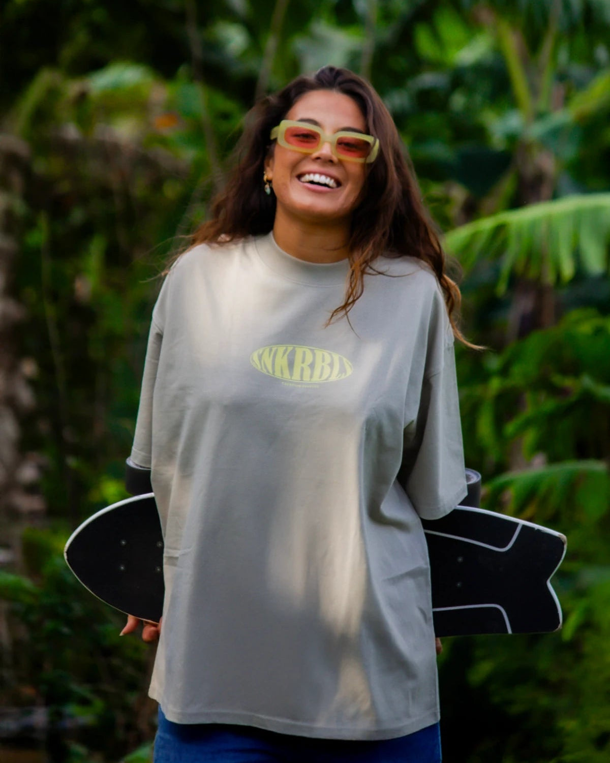 Frau im SNKRBLS Oversized T-Shirt in Stone steht lachend mit Skateboard vor tropischer Kulisse