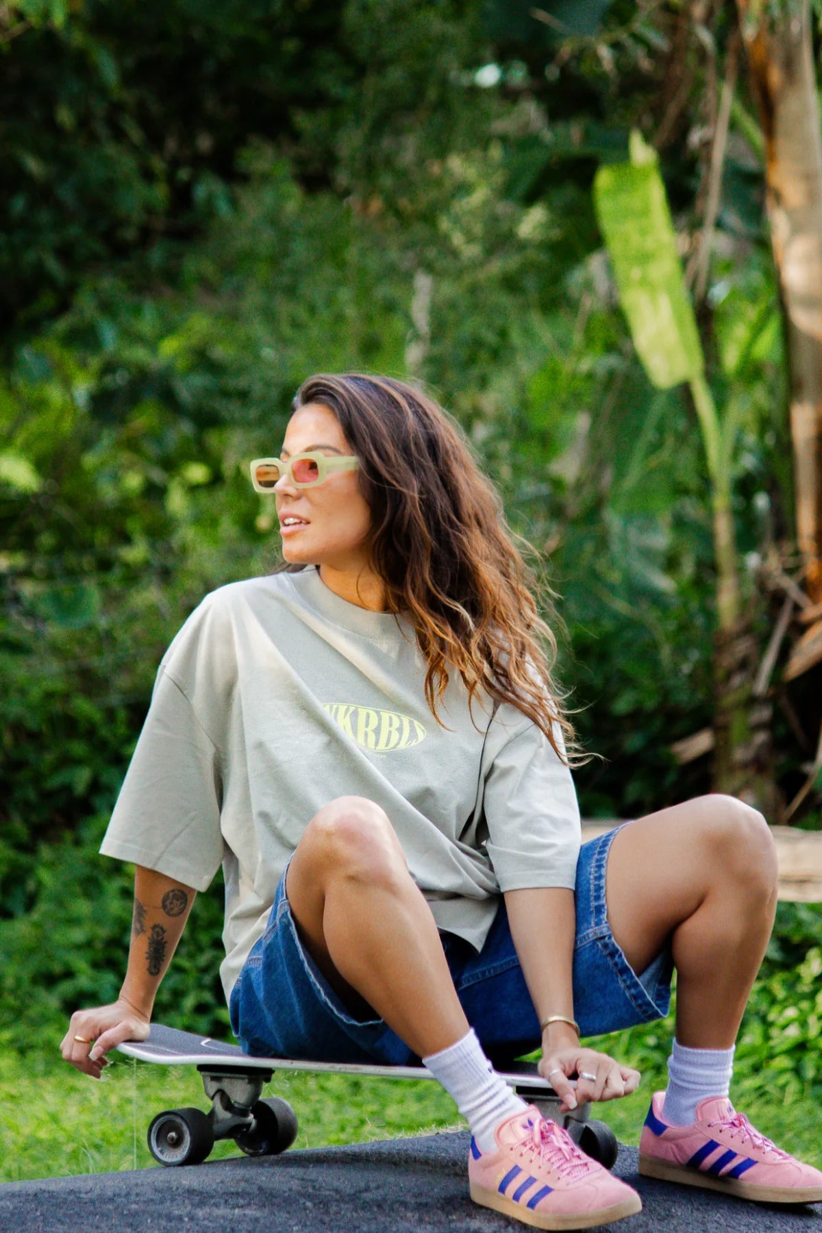 Frau im SNKRBLS Shirt Stone sitzt entspannt mit Skateboard im Grünen