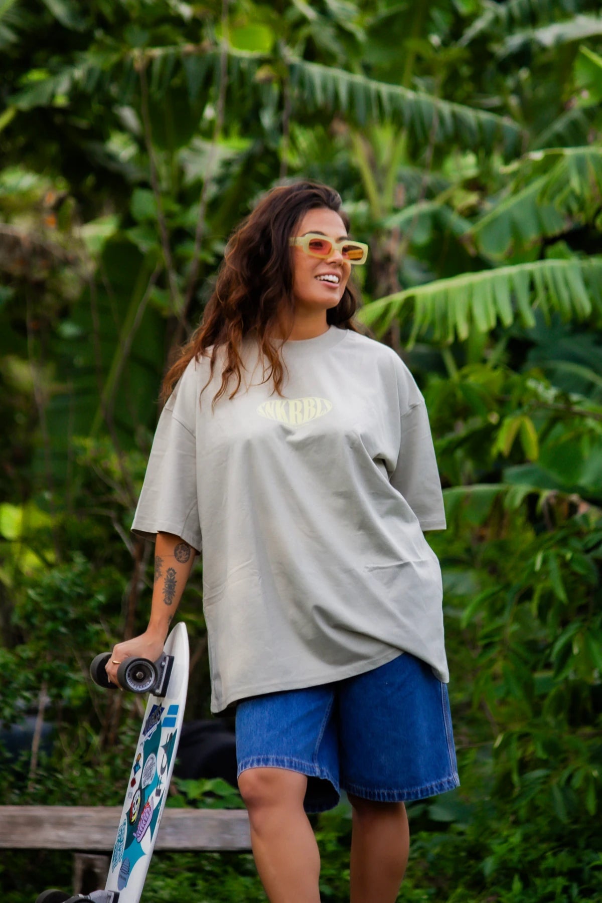Model mit SNKRBLS T-Shirt in Stone und Jeansshorts steht mit Skateboard in tropischer Umgebung