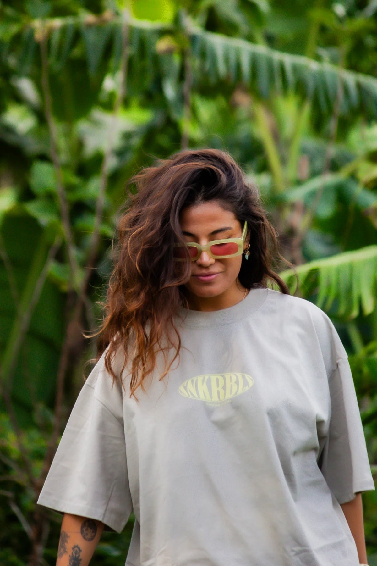 Frau mit Sonnenbrille im SNKRBLS Oversized T-Shirt Stone blickt ruhig nach unten im Grünen