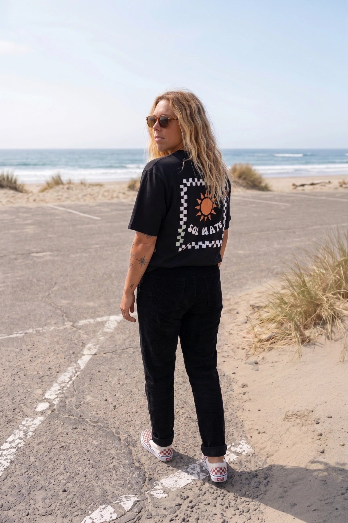 Frau trägt schwarzes Sol Mate Oversized T-Shirt von The Sunkissed Rebels mit großem Backprint am Strand