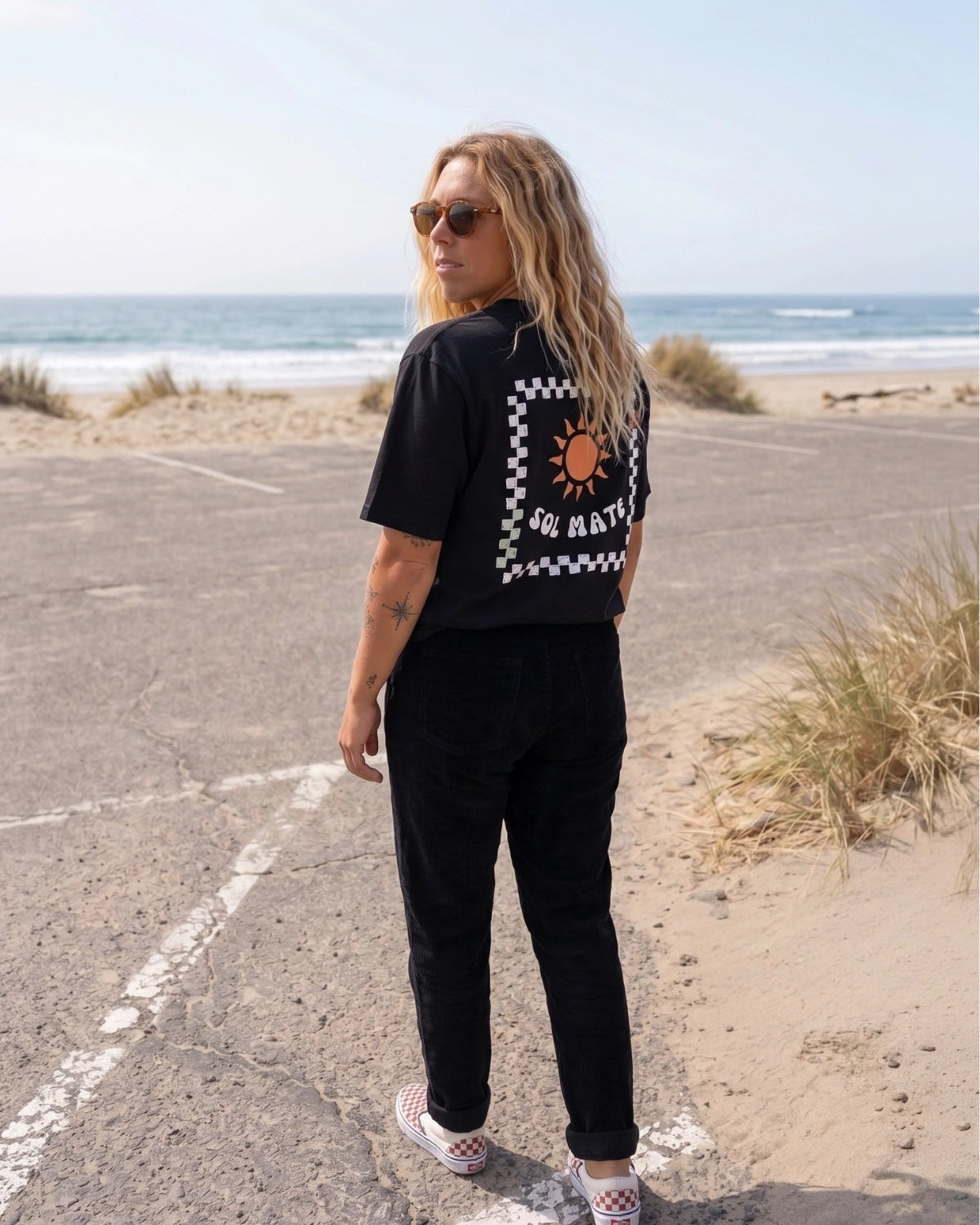 Frau trägt schwarzes Sol Mate Oversized T-Shirt von The Sunkissed Rebels mit großem Backprint am Strand