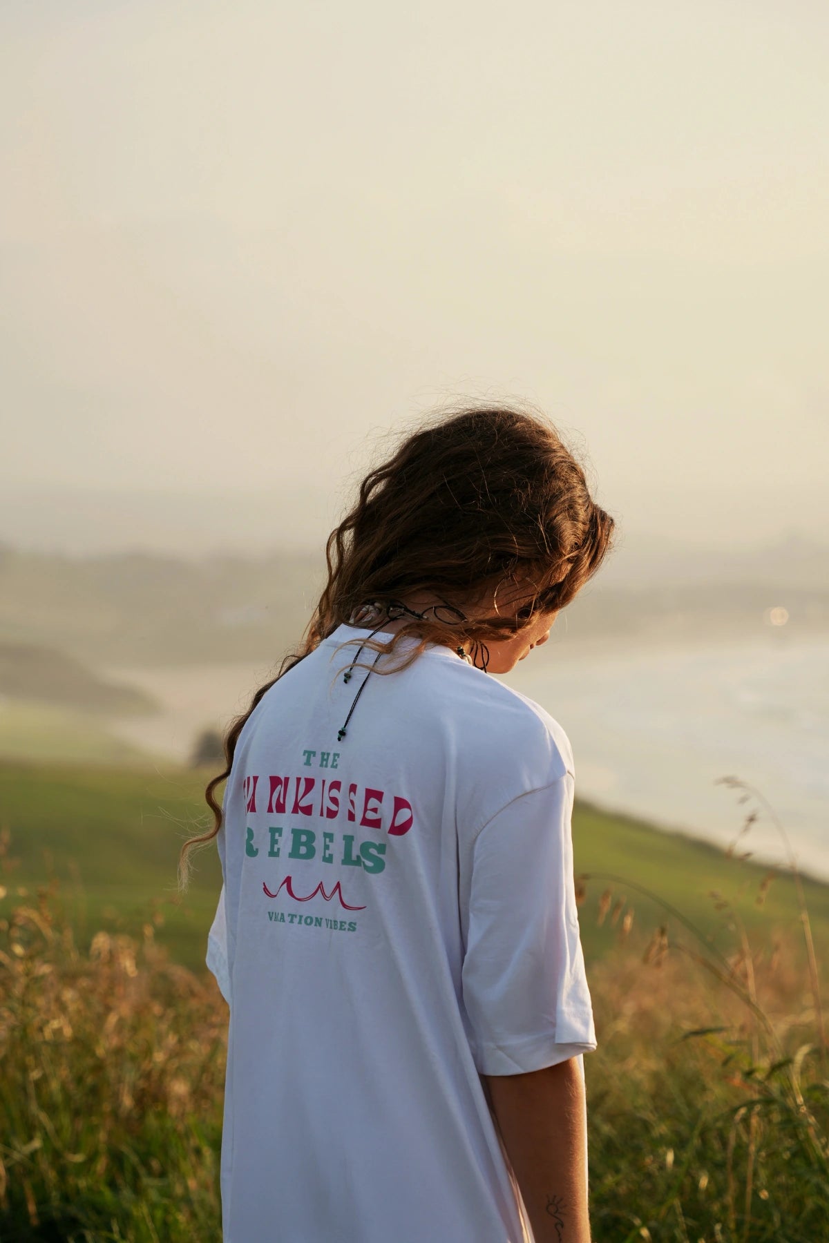 Rückansicht einer Frau mit Vacation Vibes Shirt in der goldenen Abendstimmung am Ozean