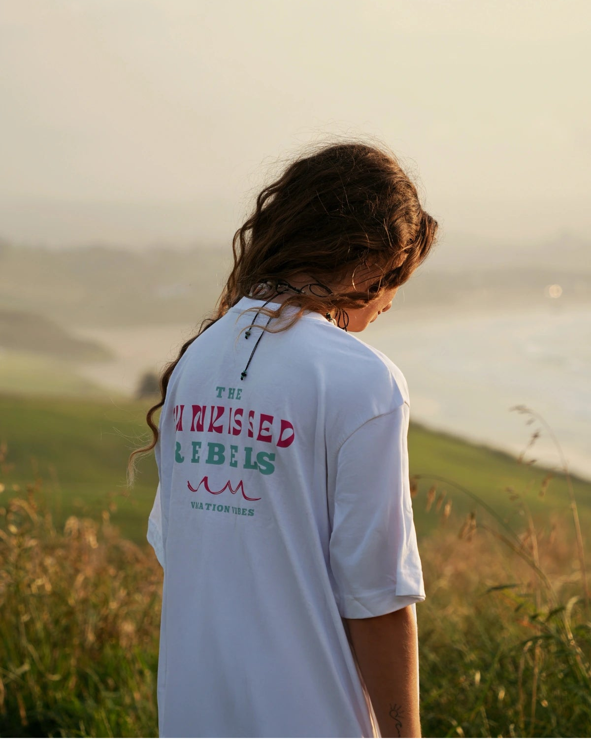 Rückansicht einer Frau mit Vacation Vibes Shirt in der goldenen Abendstimmung am Ozean