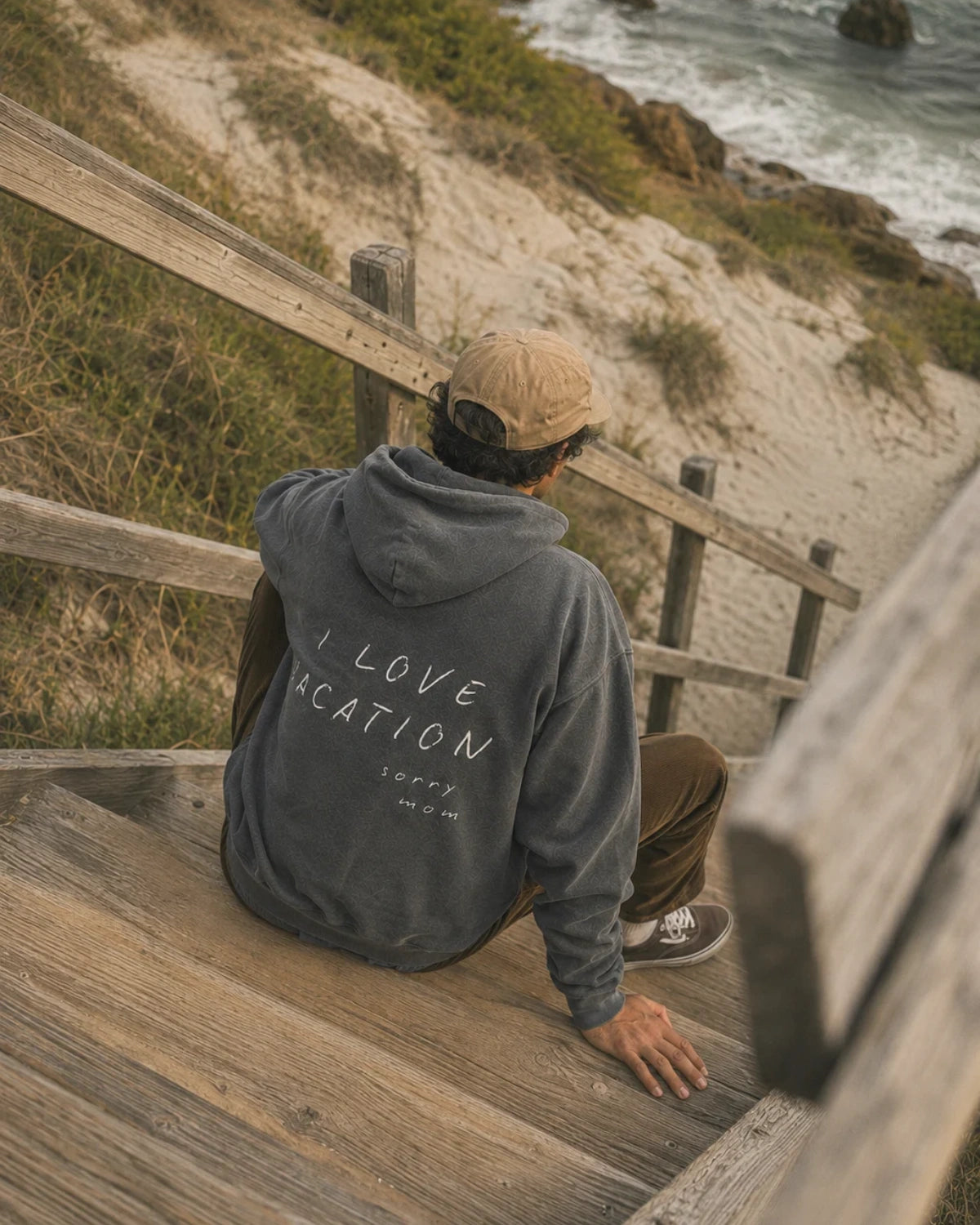 Rückansicht des Sorry Mom Oversized Hoodies mit handschriftlichem „I Love Vacation – sorry mom“ Print auf einer Holztreppe am Meer.