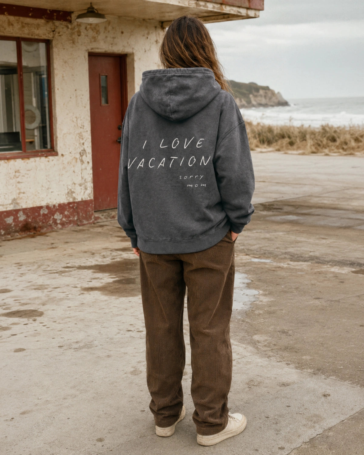 Frau im grauen Oversized Hoodie von The Sunkissed Rebels mit „I Love Vacation – sorry mom“ Backprint vor einem alten Küstenhaus.