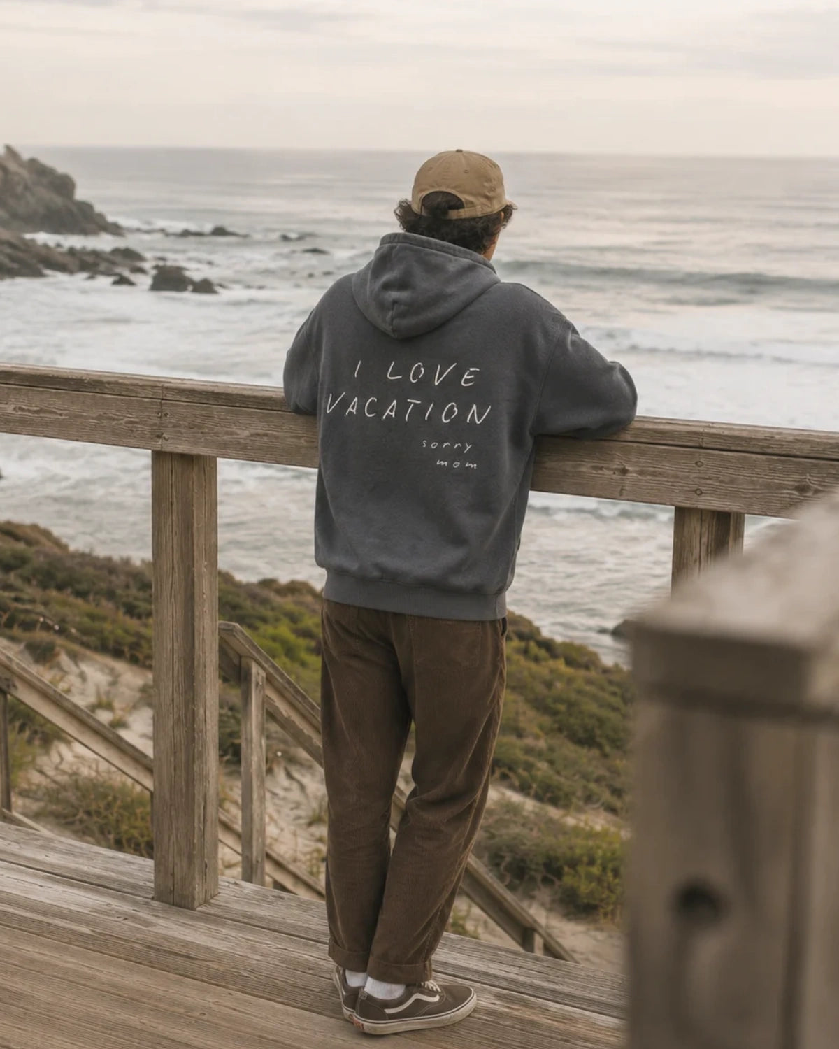 Oversized Hoodie in verwaschenem Anthrazit mit weißem Vacation-Backprint, getragen von einem Mann am Aussichtspunkt über dem Meer.