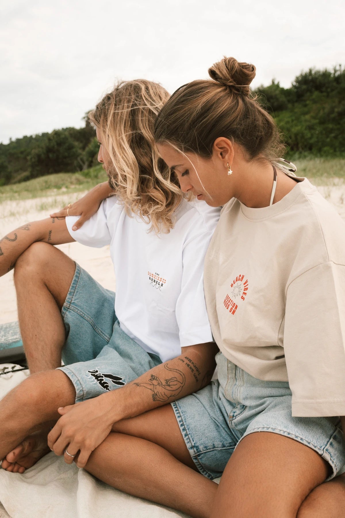 Strandaufnahme von zwei Models in Sunkissed Rebels Shirts beim Sitzen im Sand.