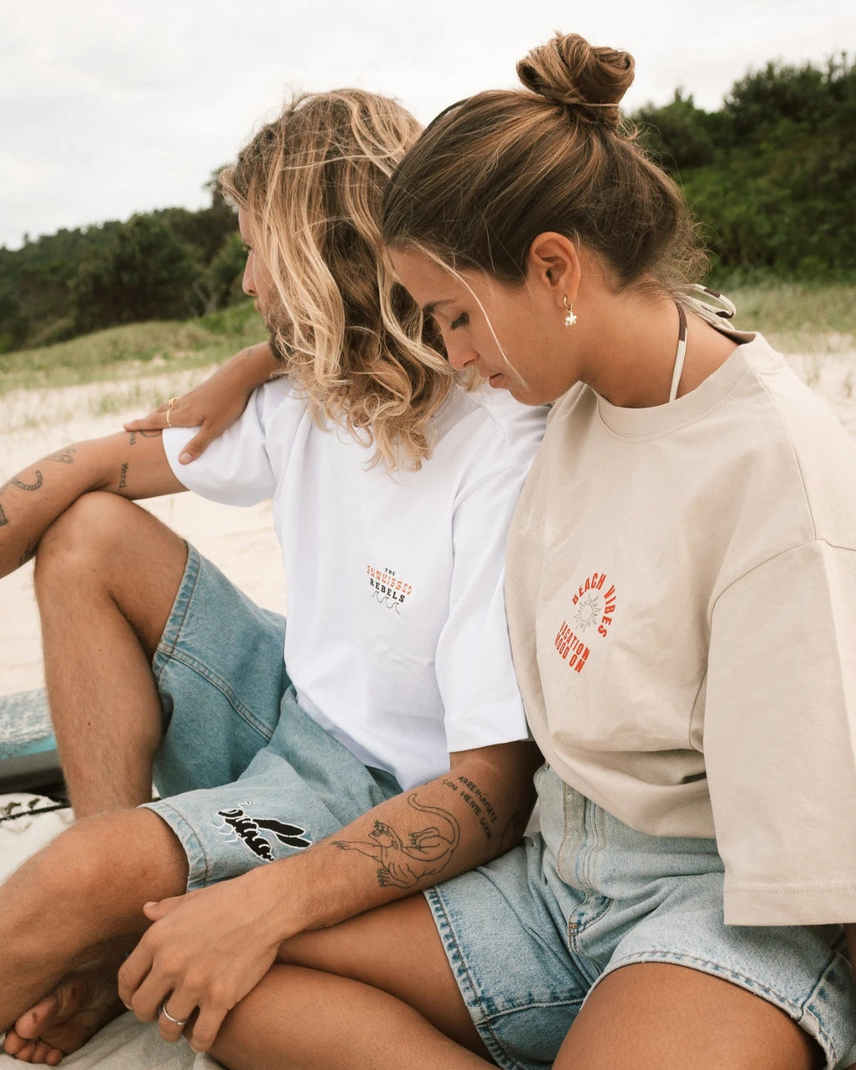 Strandaufnahme von zwei Models in Sunkissed Rebels Shirts beim Sitzen im Sand.
