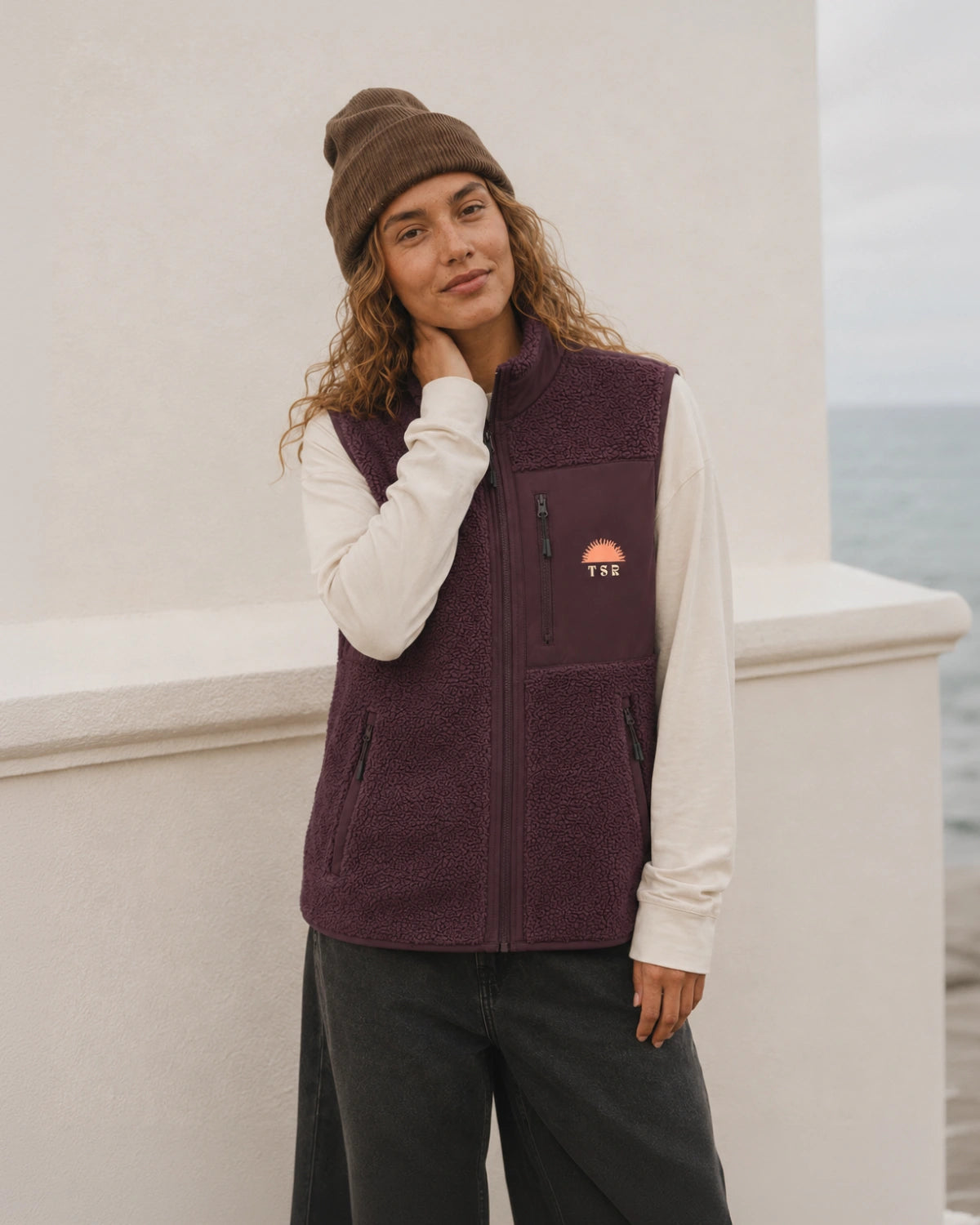 Frau in der Sundown Fleece Vest in Burgundy mit weicher Sherpa-Struktur, TSR-Sonnenlogo und brauner Beanie vor einer hellen Hauswand.