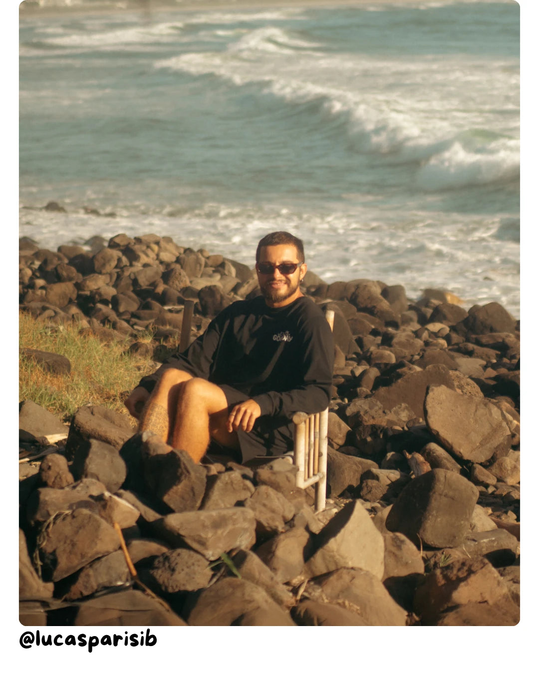 Model von The Sunkissed Rebels sitzt entspannt auf Felsen am Meer und trägt einen schwarzen Hoodie.
