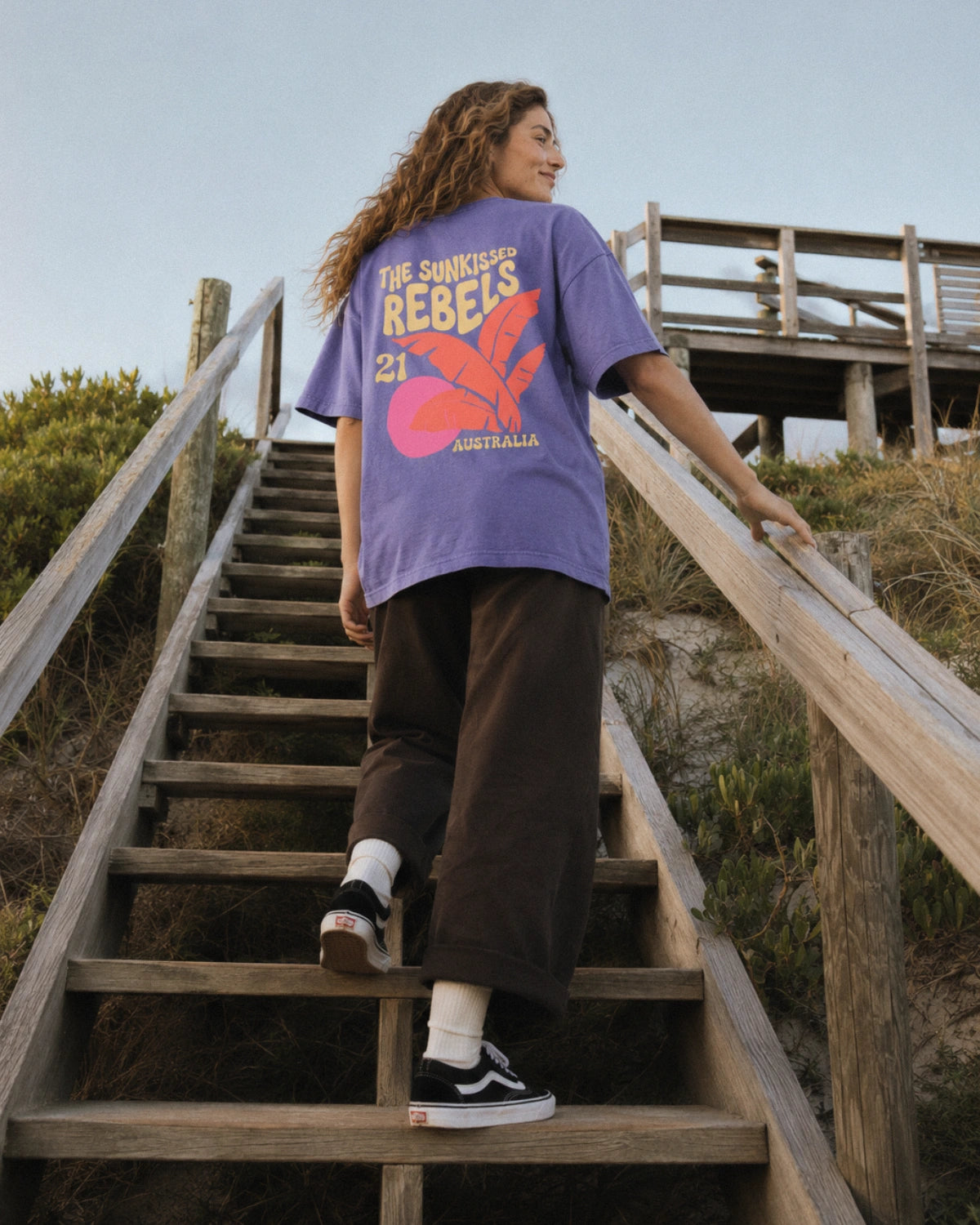 Frau im violetten Oversized Heavy T-Shirt von The Sunkissed Rebels mit großem Australia-Palmenprint auf einer Holztreppe in den Dünen.