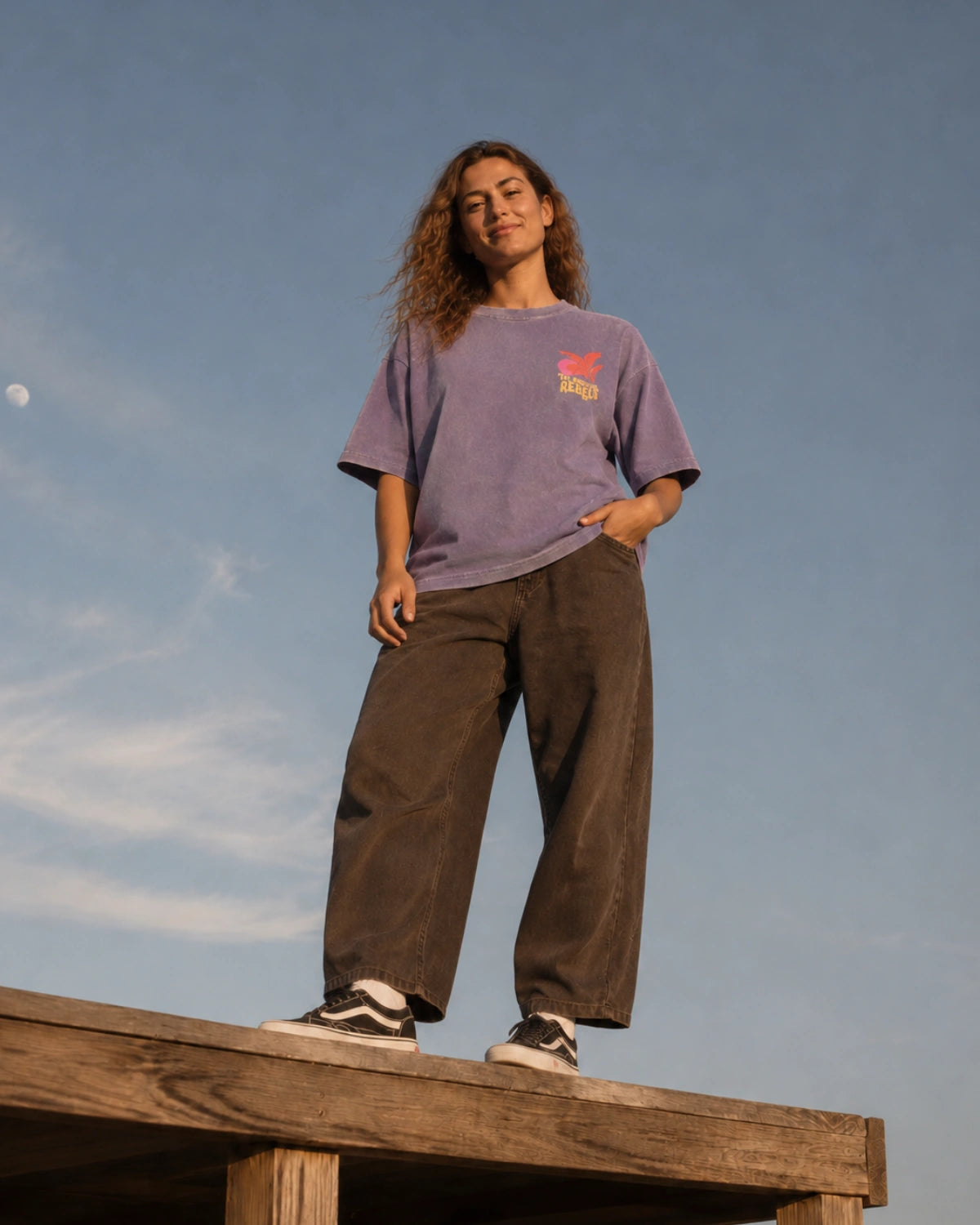 Frau trägt das Sunkissed Rebels Heavy Tee in Vintage Violet mit lockerem Oversized Fit, kombiniert mit brauner Hose und Sneakers.