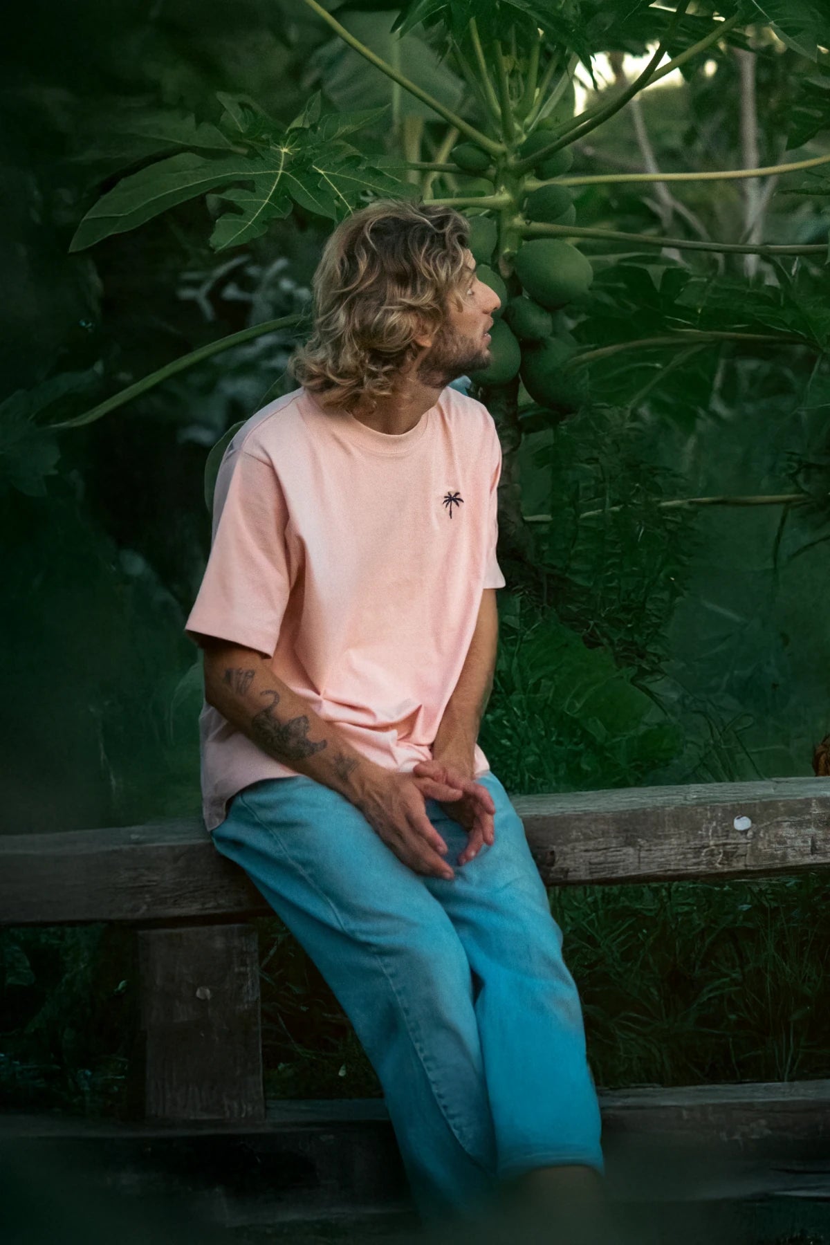 Model mit coral Oversized T-Shirt von The Sunkissed Rebels sitzt auf Holzbank in tropischer Kulisse