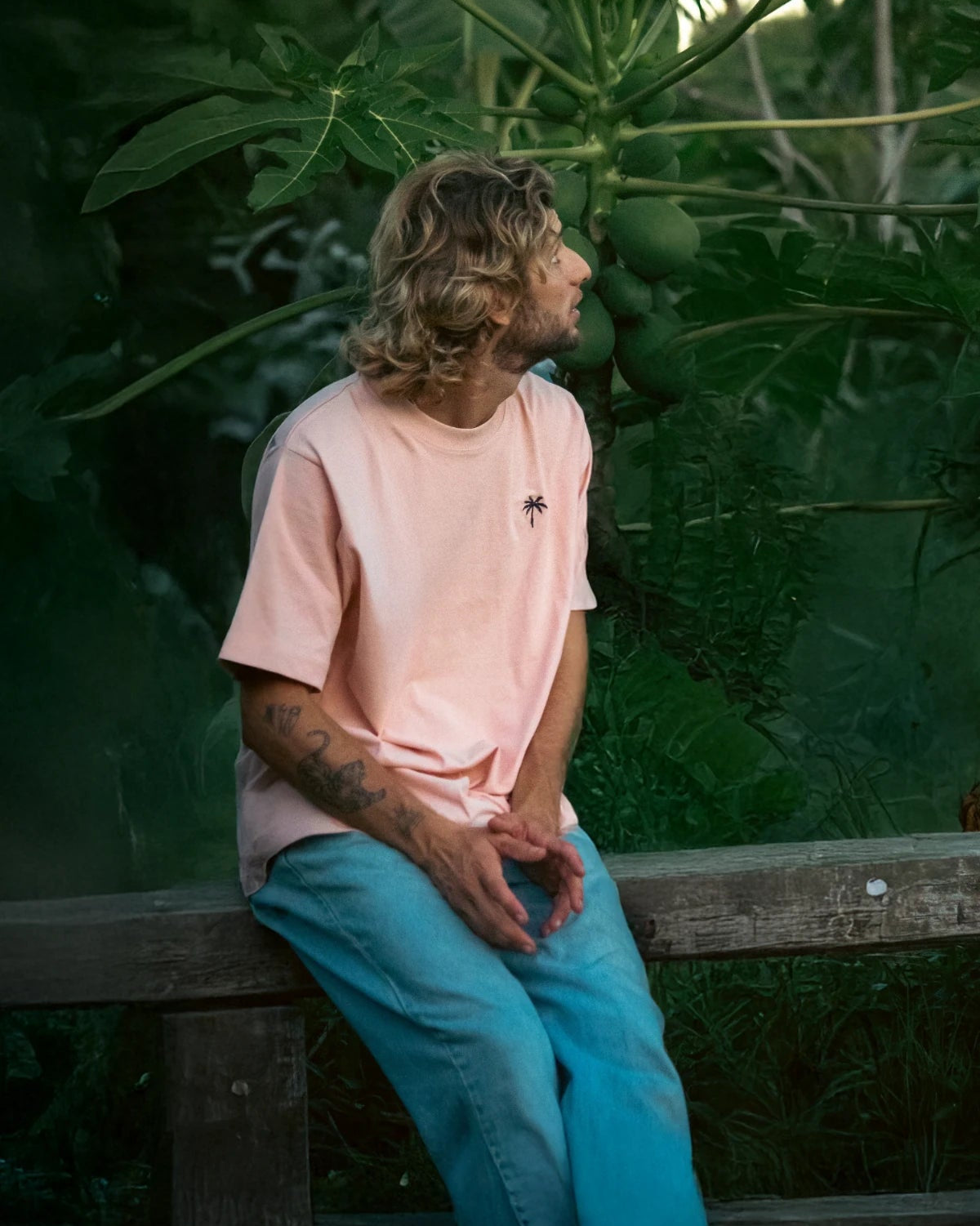 Model mit coral Oversized T-Shirt von The Sunkissed Rebels sitzt auf Holzbank in tropischer Kulisse