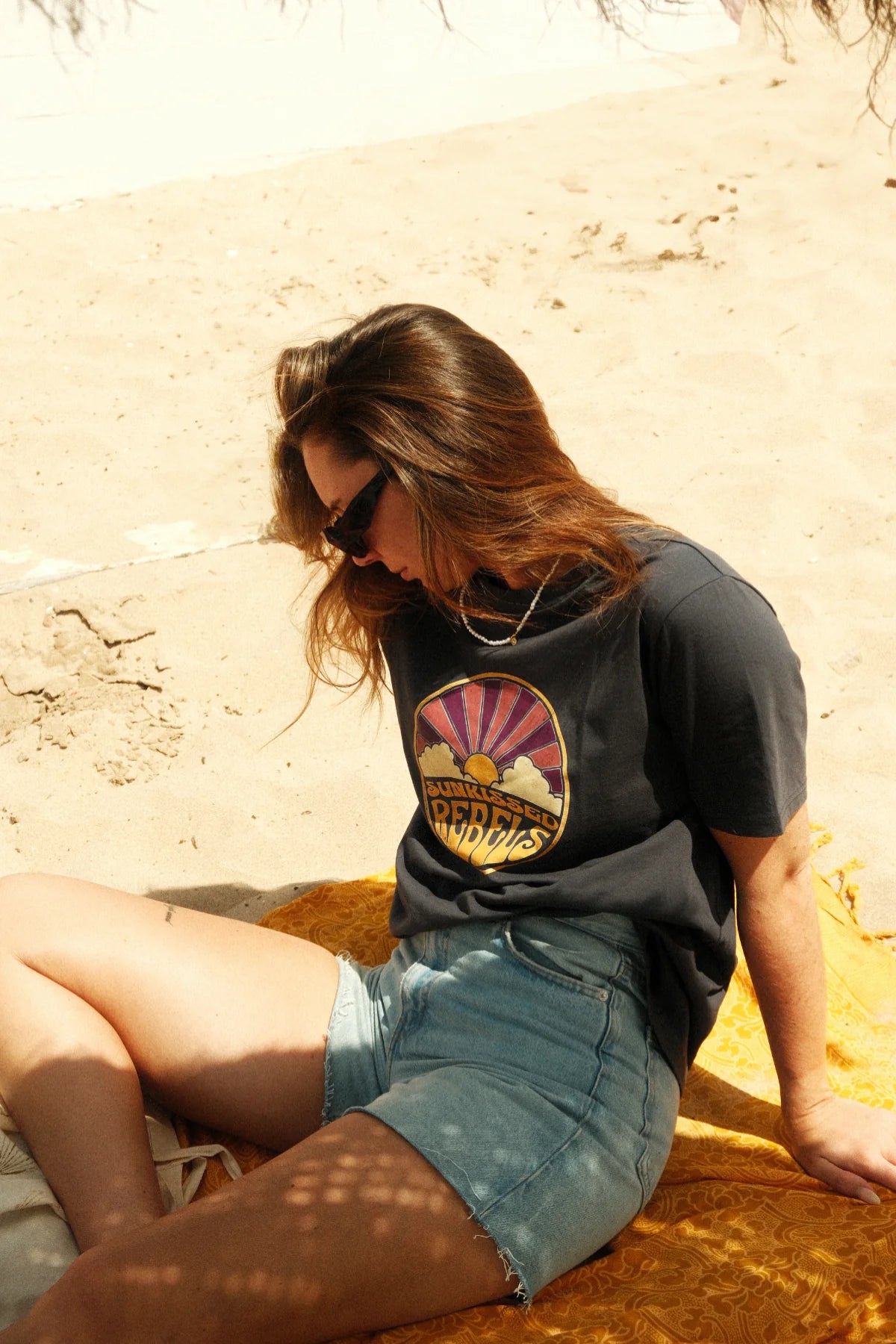 Frau trägt Sunkissed Rebels T-Shirt in Dunkelgrau und sitzt entspannt auf Strandtuch im Sand.