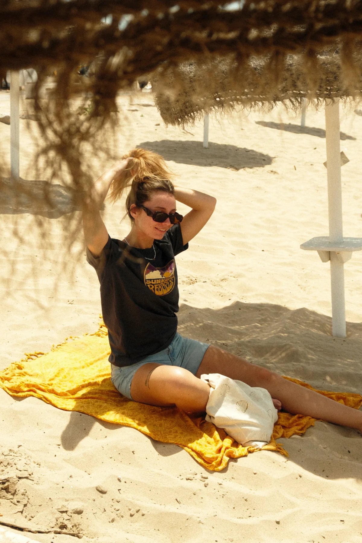 Model bindet ihre Haare zusammen und trägt das Sunkissed Rebels T-Shirt sitzend unter Palmen-Schatten.